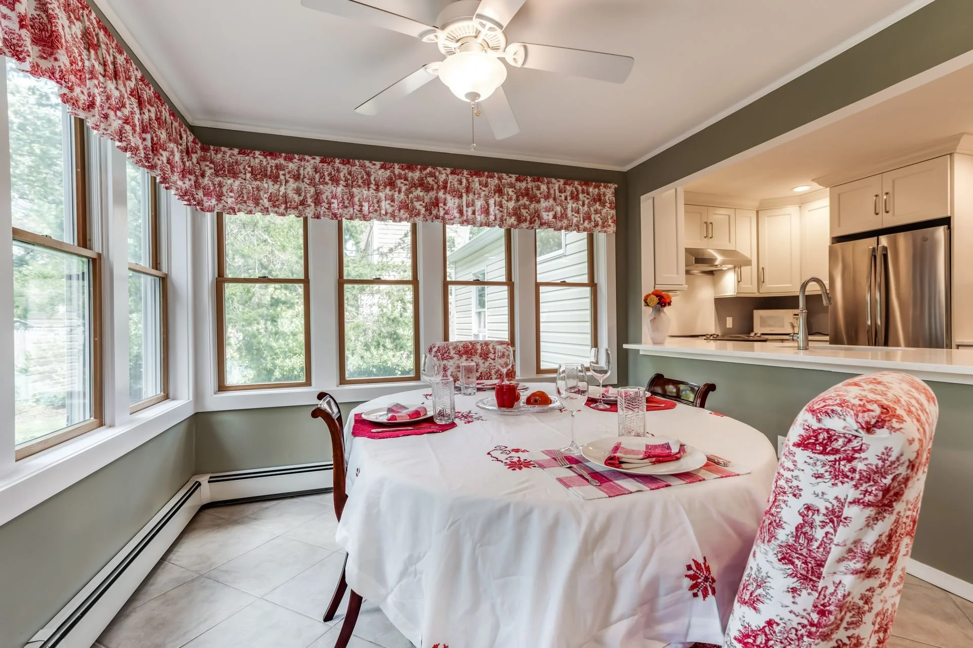 A dining area with a round table covered with a white tablecloth decorated with red embroidery, set with glassware, plates, and a red apple. Surrounding chairs are upholstered with red and white toile fabric, matching the red floral curtain valance o