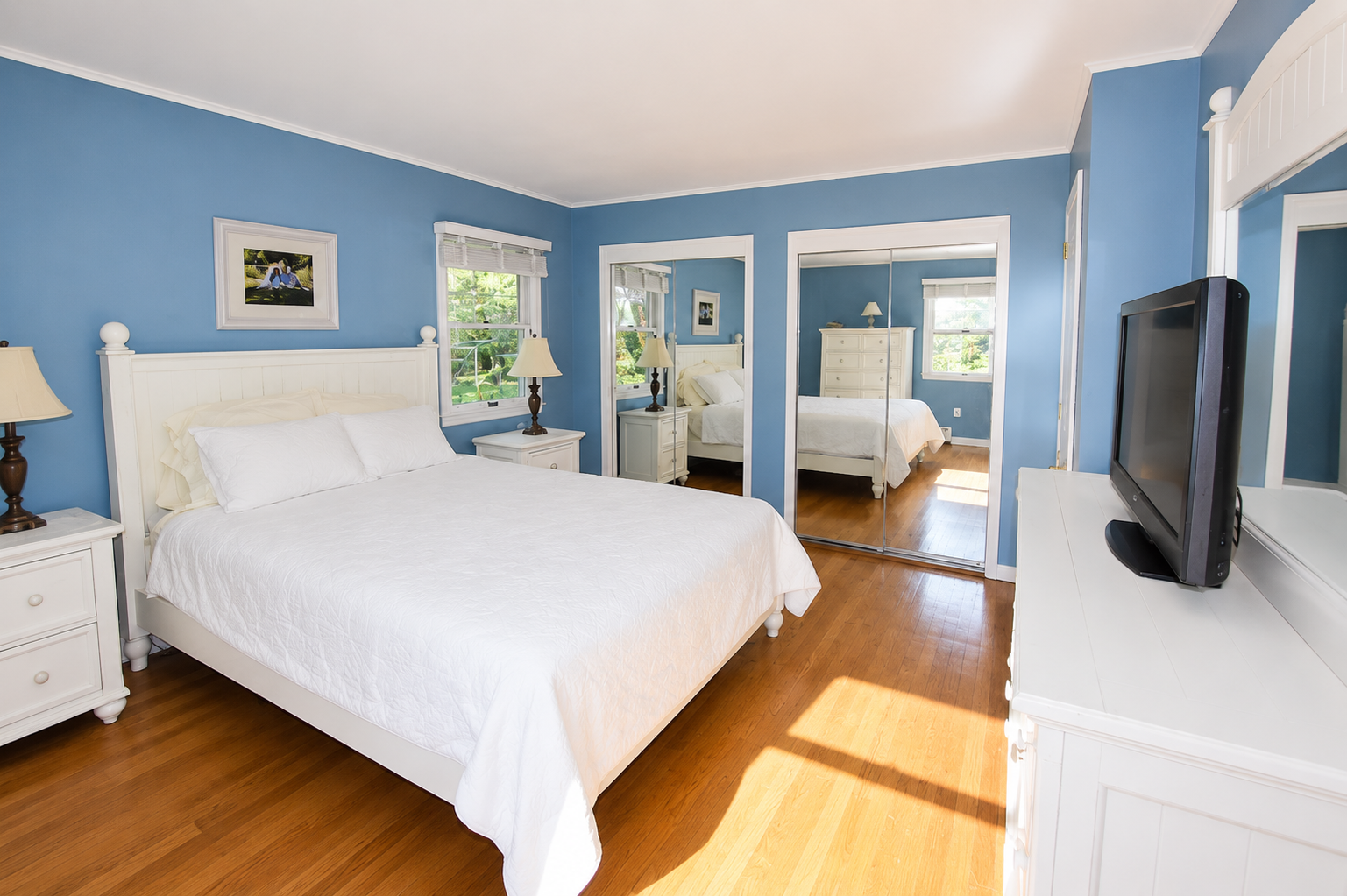 Bedroom with blue walls, white bed with white bedding, two white nightstands with lamps, a dresser with a mirror, a TV on a white dresser, and large mirrored closet doors