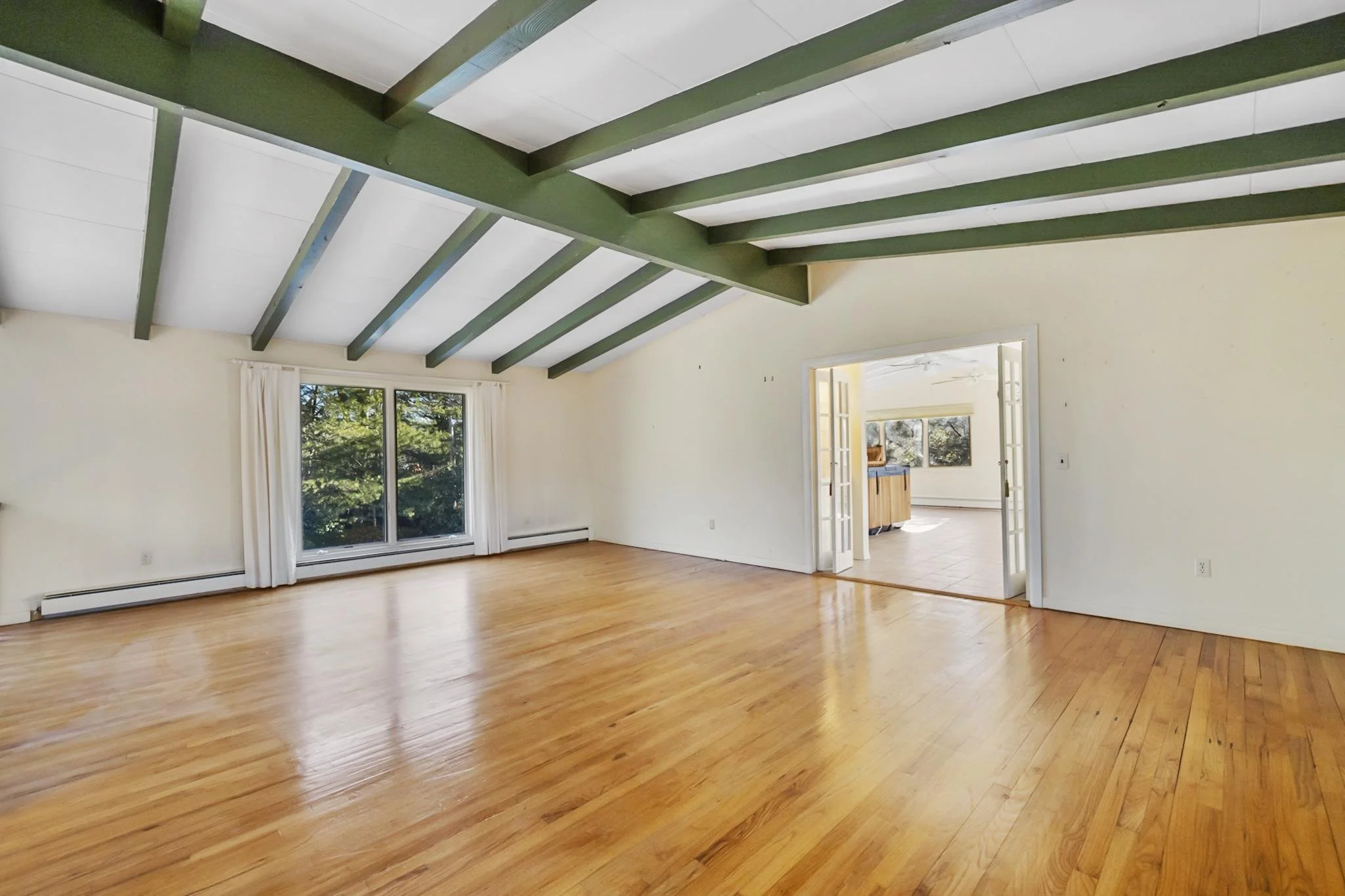 A spacious empty living room with hardwood floors, large windows with white curtains, and an open doorway leading to another room with visible cabinets and windows. The ceiling has exposed green beams and white panels.