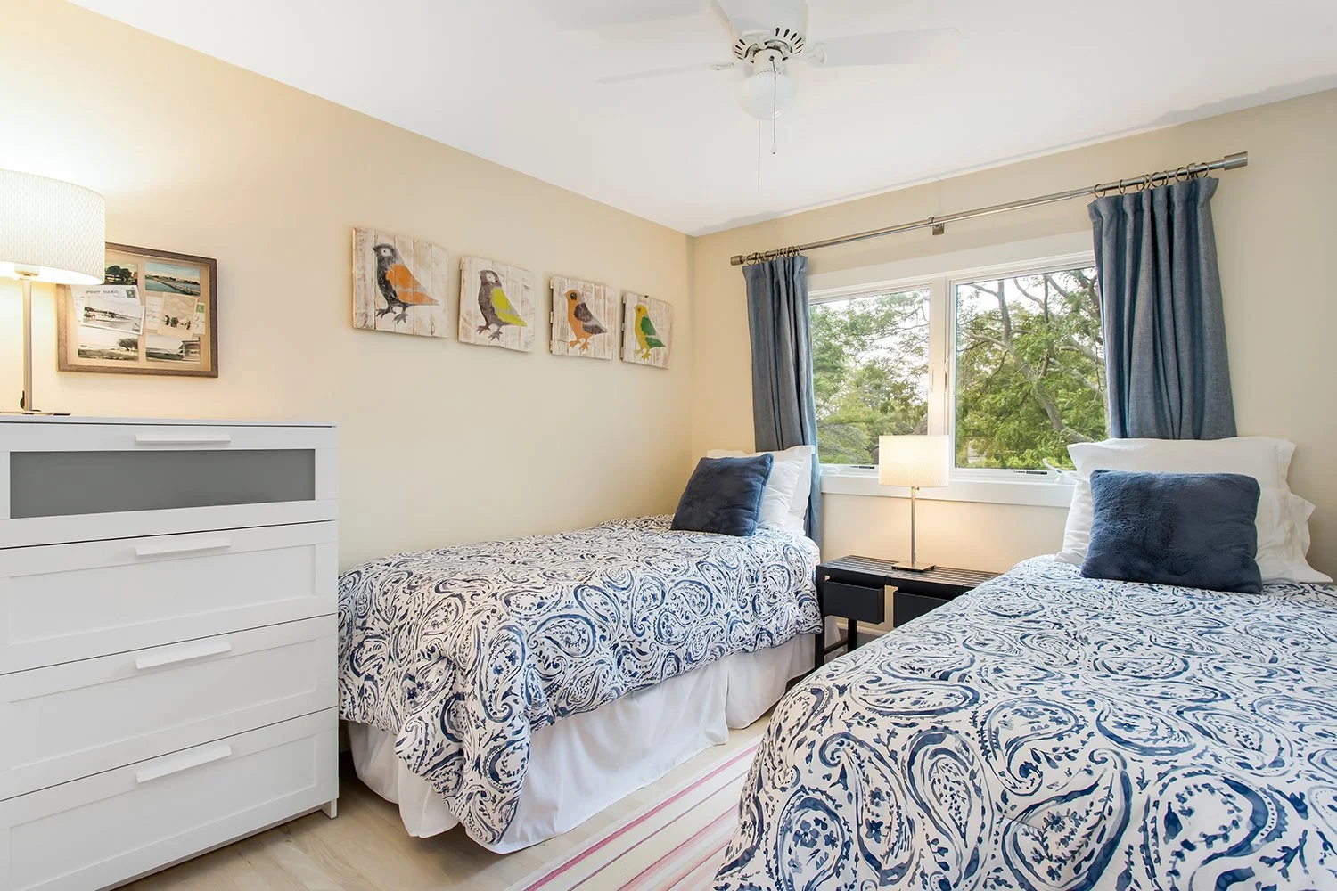 Bedroom with two twin beds with white and blue patterned bedding, dark blue pillows, two windows with blue curtains, a white dresser, and wall art of colorful bird paintings.
