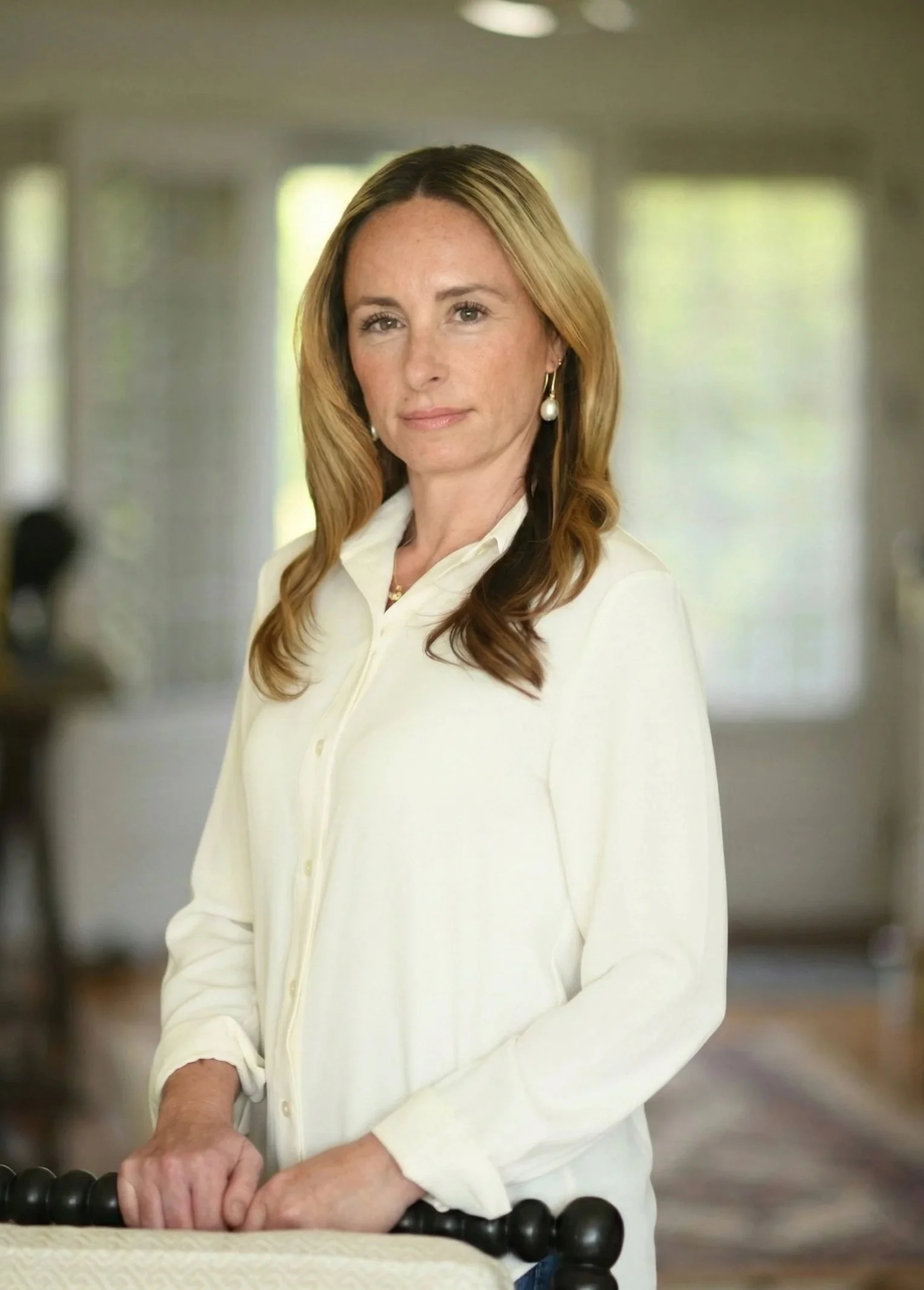 A middle-aged woman with light skin and red hair, wearing pearl earrings and a cream-colored blouse, standing indoors with a blurred background and her hands resting on a black bead railing.