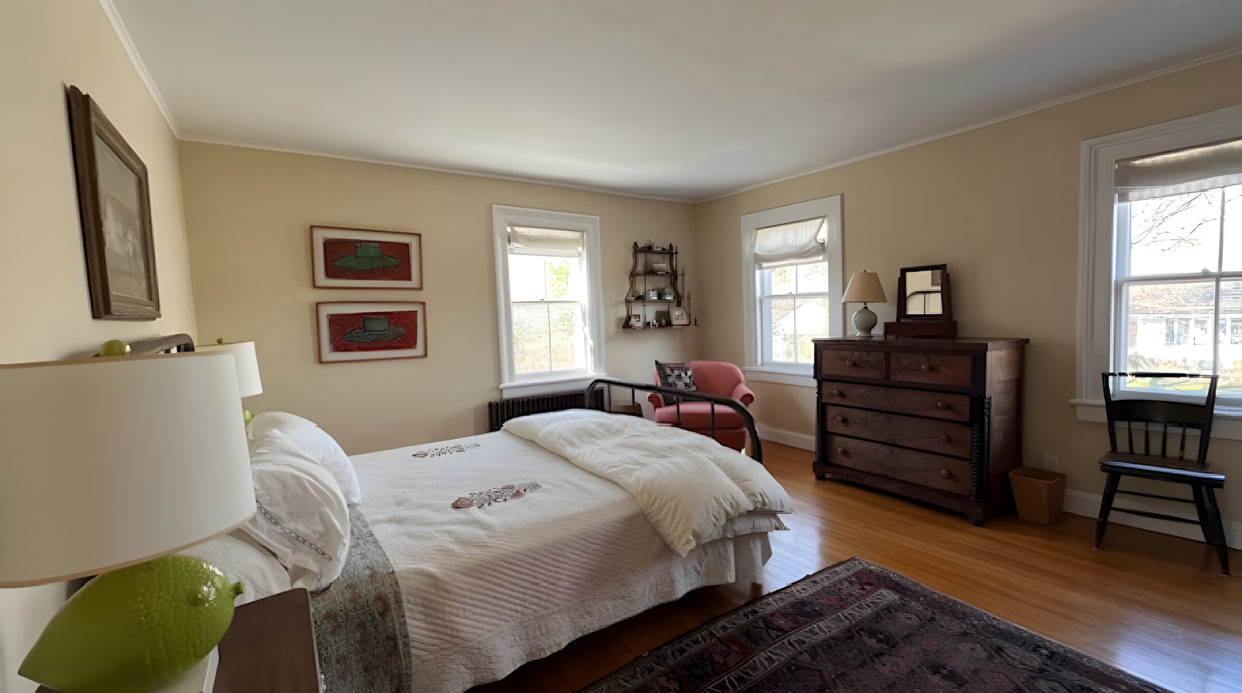 A cozy bedroom with a bed, framed artwork, a pink armchair, a wooden dresser, and several windows with white blinds, allowing natural light into the room.