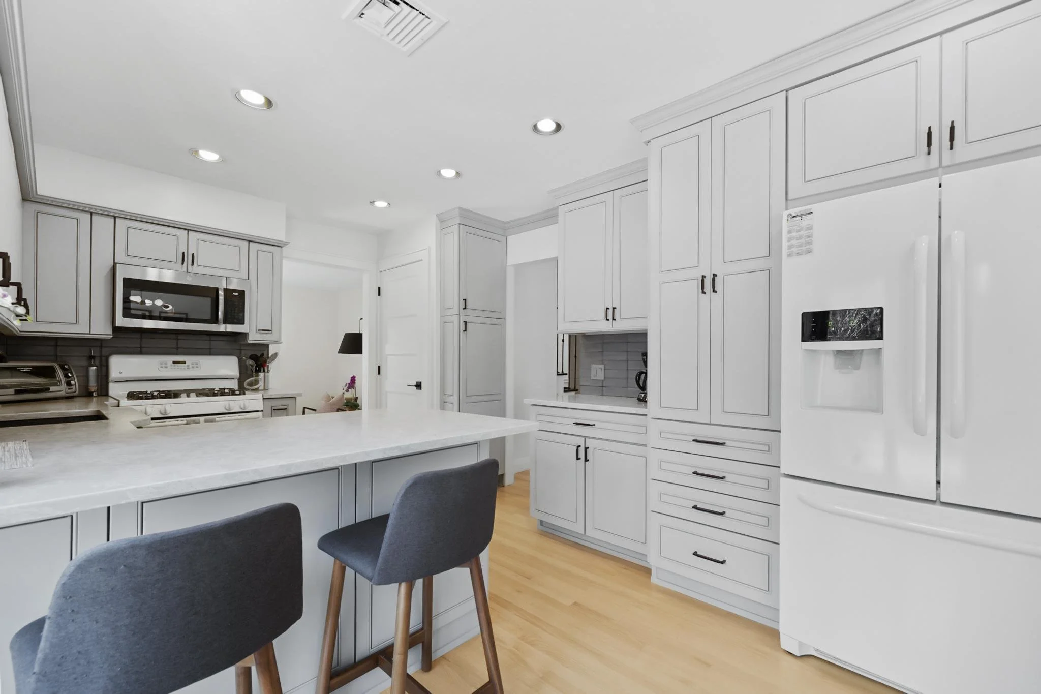 A modern white kitchen with white cabinets, a white island with a marble countertop, black hardware, and stainless steel appliances. Two dark gray chairs are at the island, and the floor is wooden. Recessed ceiling lights and a smoke detector are vis