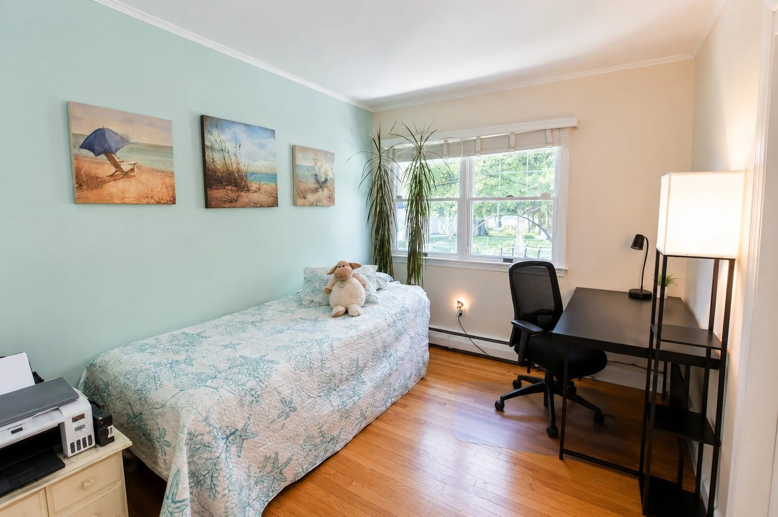 A bedroom with light green and white walls, a window with blinds, hardwood floors, a bed with a stuffed animal, a desk with a lamp, an office chair, and wall art of beach scenes.