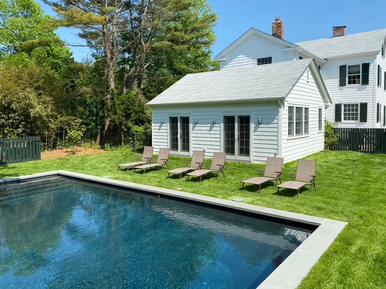 Backyard with swimming pool, six lounge chairs, white house with black shutters, and surrounded by green trees and grass.