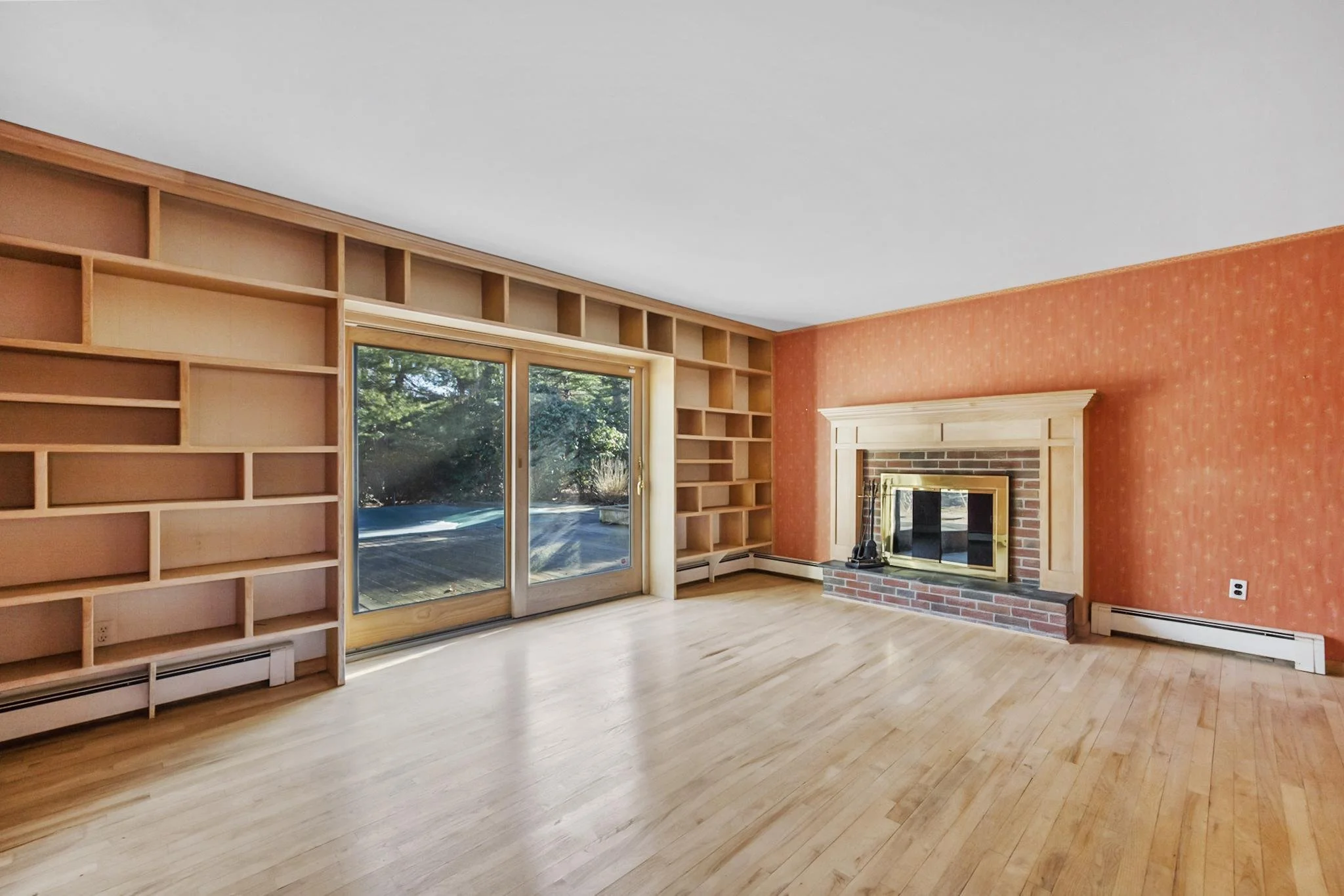 Living room with floor-to-ceiling wooden bookshelves, hardwood floors, large window showing greenery outside, a brick fireplace with wooden mantel, and an orange patterned wall.
