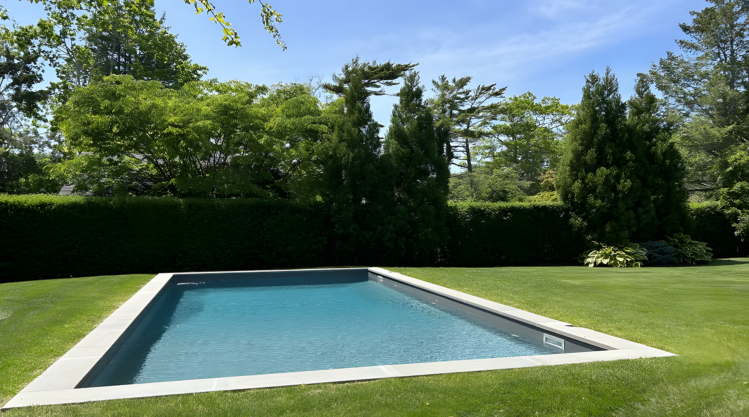 A rectangular backyard pool surrounded by green grass and tall trees, under a partly cloudy sky.