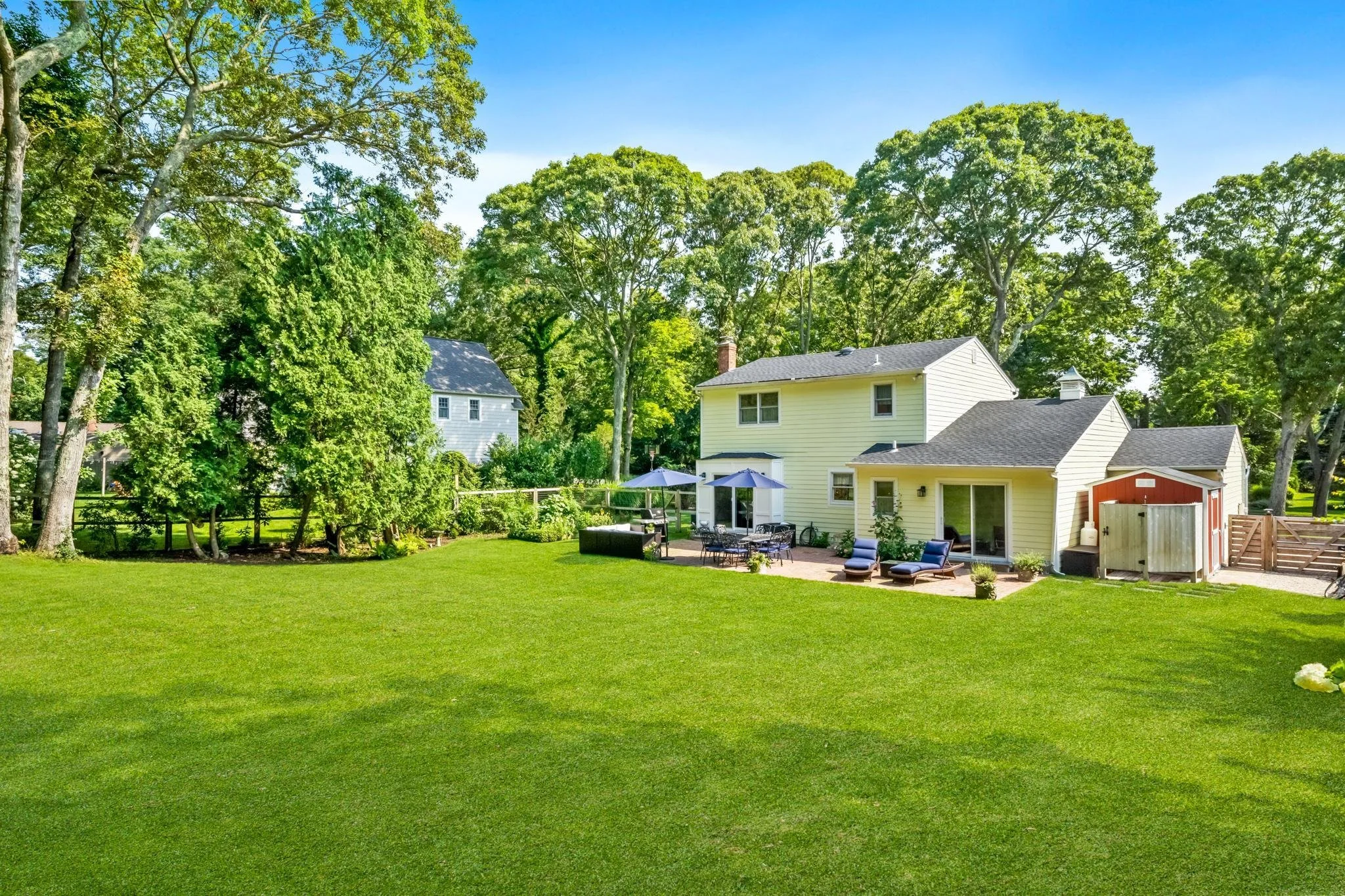 Backyard with green grass, trees, and a white house with a black roof. Outdoor furniture including chairs, a table with umbrellas, and lounge chairs are on a patio.