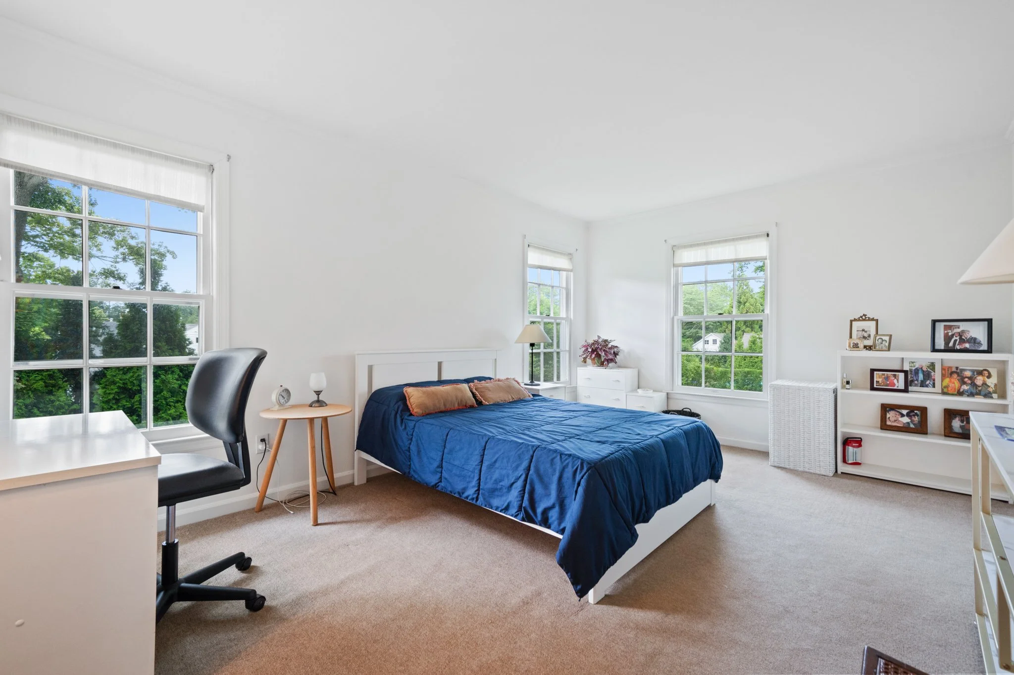 A bright bedroom with three large windows showing green trees outside. The room has a white bed frame with a blue bedspread and brown pillows. There is a nightstand with a lamp and potted plant. A white bookshelf with framed photos and a small basket