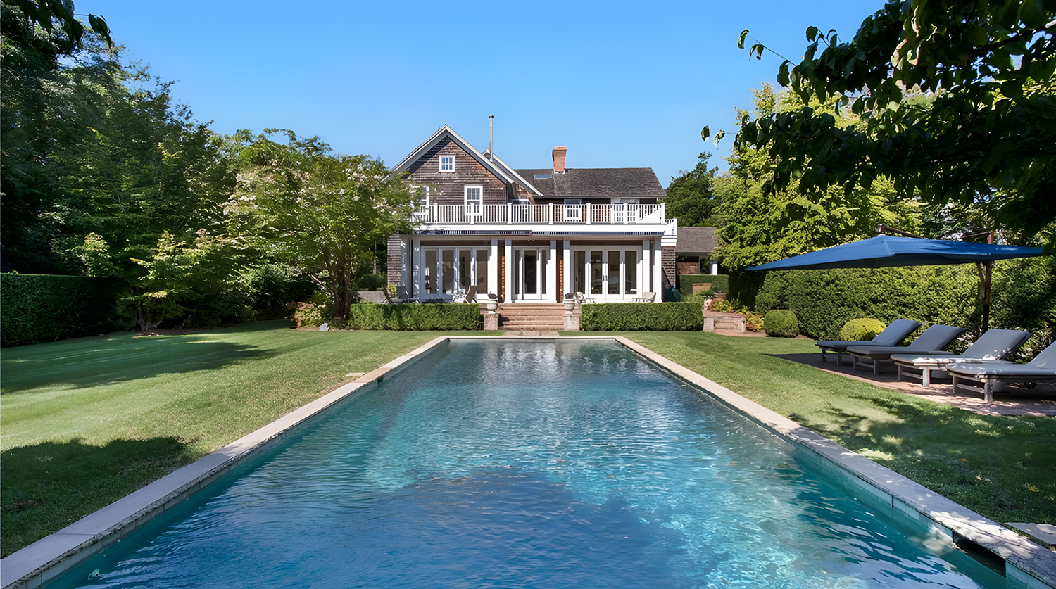 A backyard with a rectangular swimming pool, green lawn, and trees. There is a house in the background with a porch and patio area, along with lounge chairs and an umbrella on the right side.