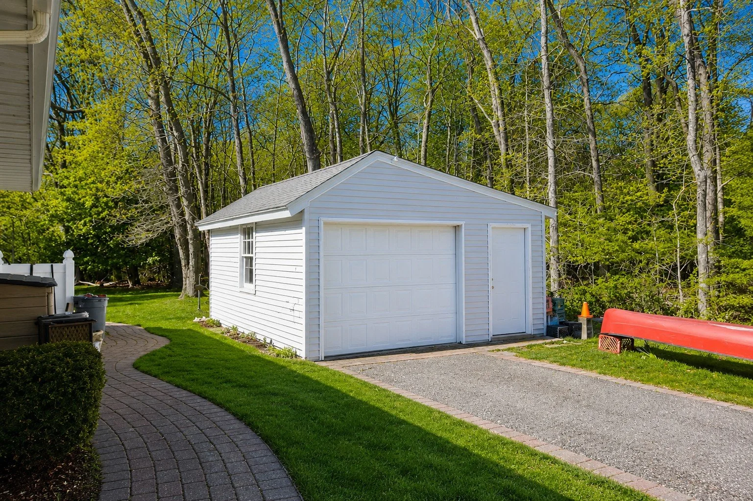 White garage with a gray roof situated in a backyard surrounded by green trees and grass, with a paved driveway and a curved brick pathway leading to the garage.