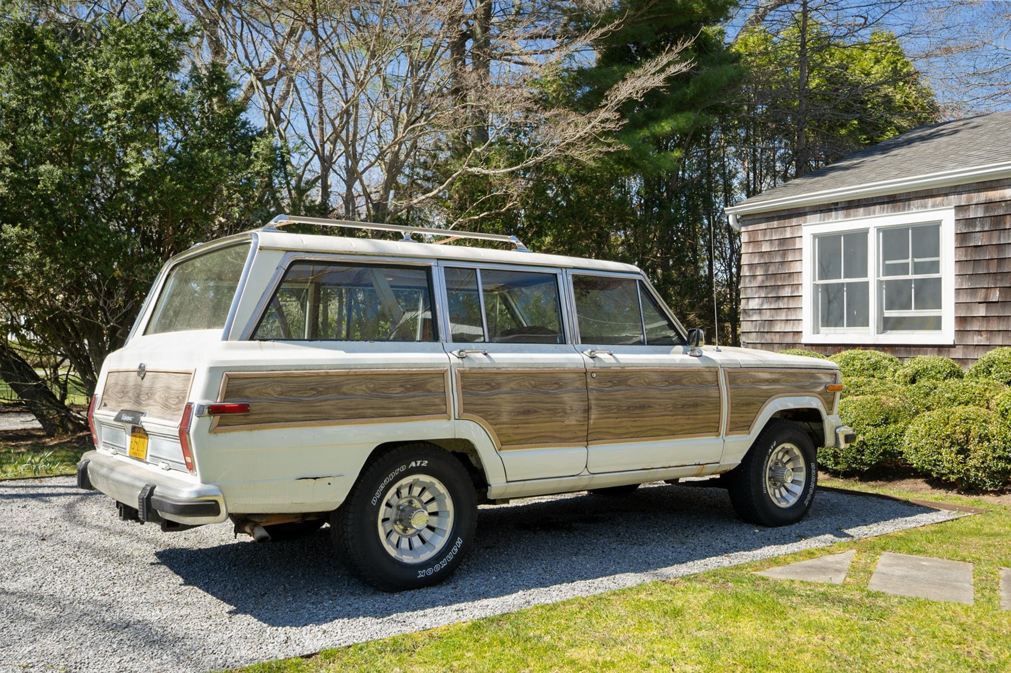 A vintage station wagon with wood paneling parked on a gravel driveway with grass. There is a house with wooden shingles and white-framed windows on the right and a background of trees with budding and bare branches under a blue sky.