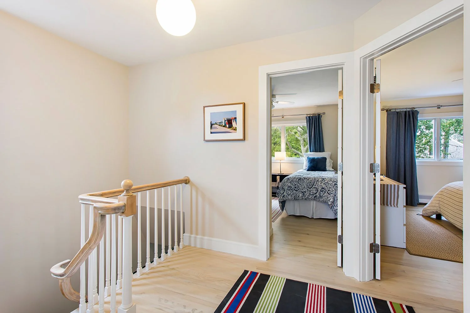 View of a hallway with two open doors leading to bedrooms, featuring framed art on the wall, a security-style striped rug, and a staircase with a wooden handrail.