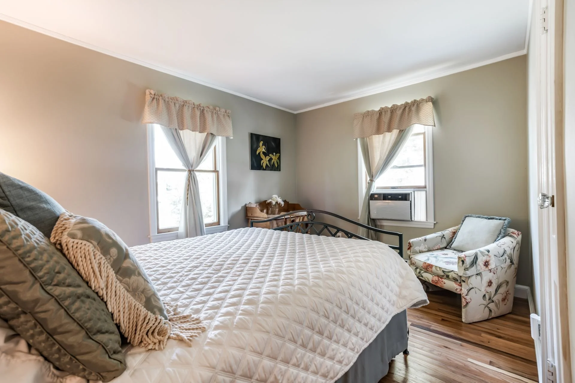 A cozy bedroom with a large bed, floral armchair, and windows with curtains, heater, and wooden flooring.
