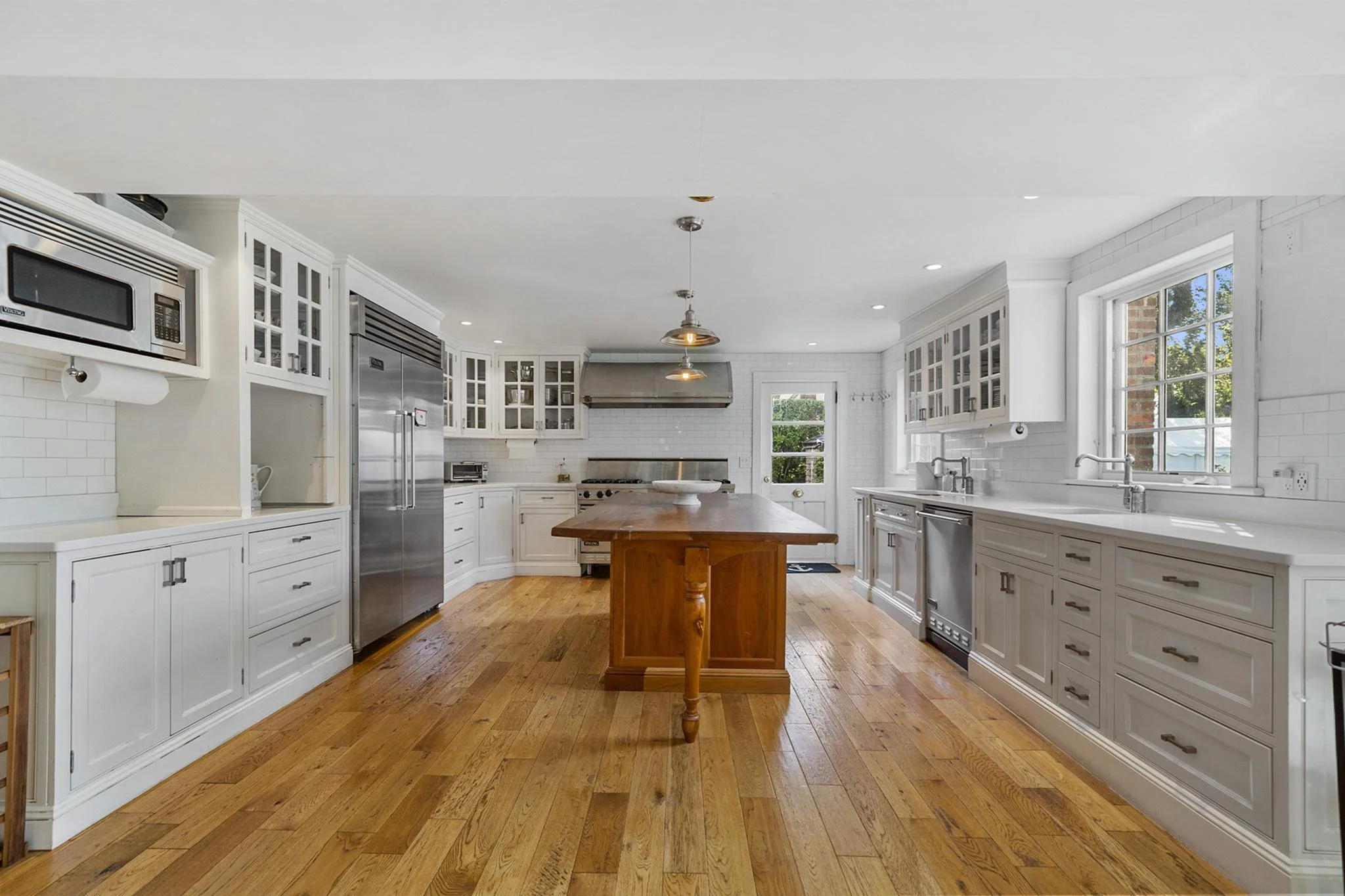 Bright white kitchen with wooden floor, white cabinets with glass doors, stainless steel appliances, and large windows with a view of trees outside.