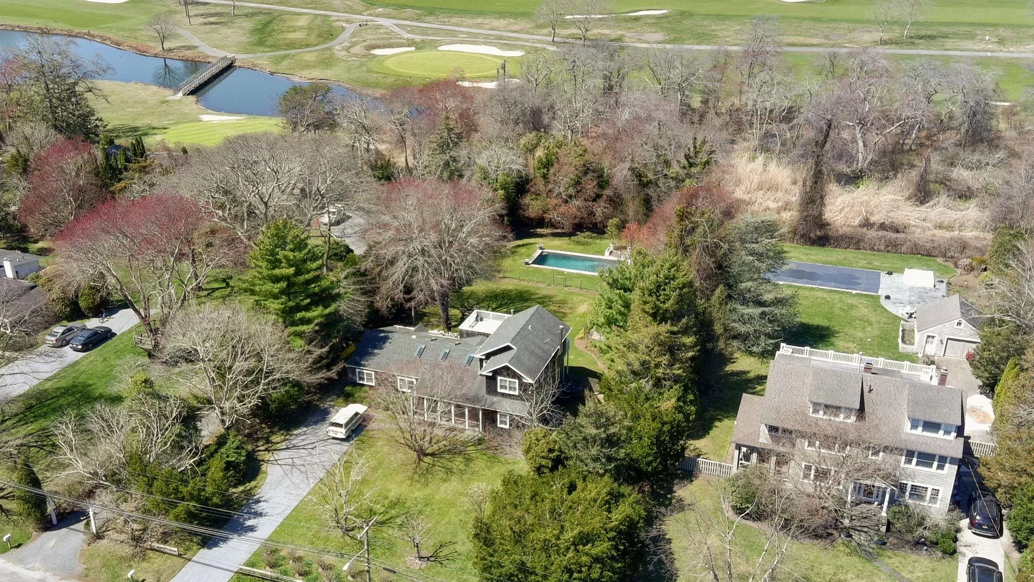 An aerial view of a suburban neighborhood with houses, trees, a pond, and a golf course in the background.