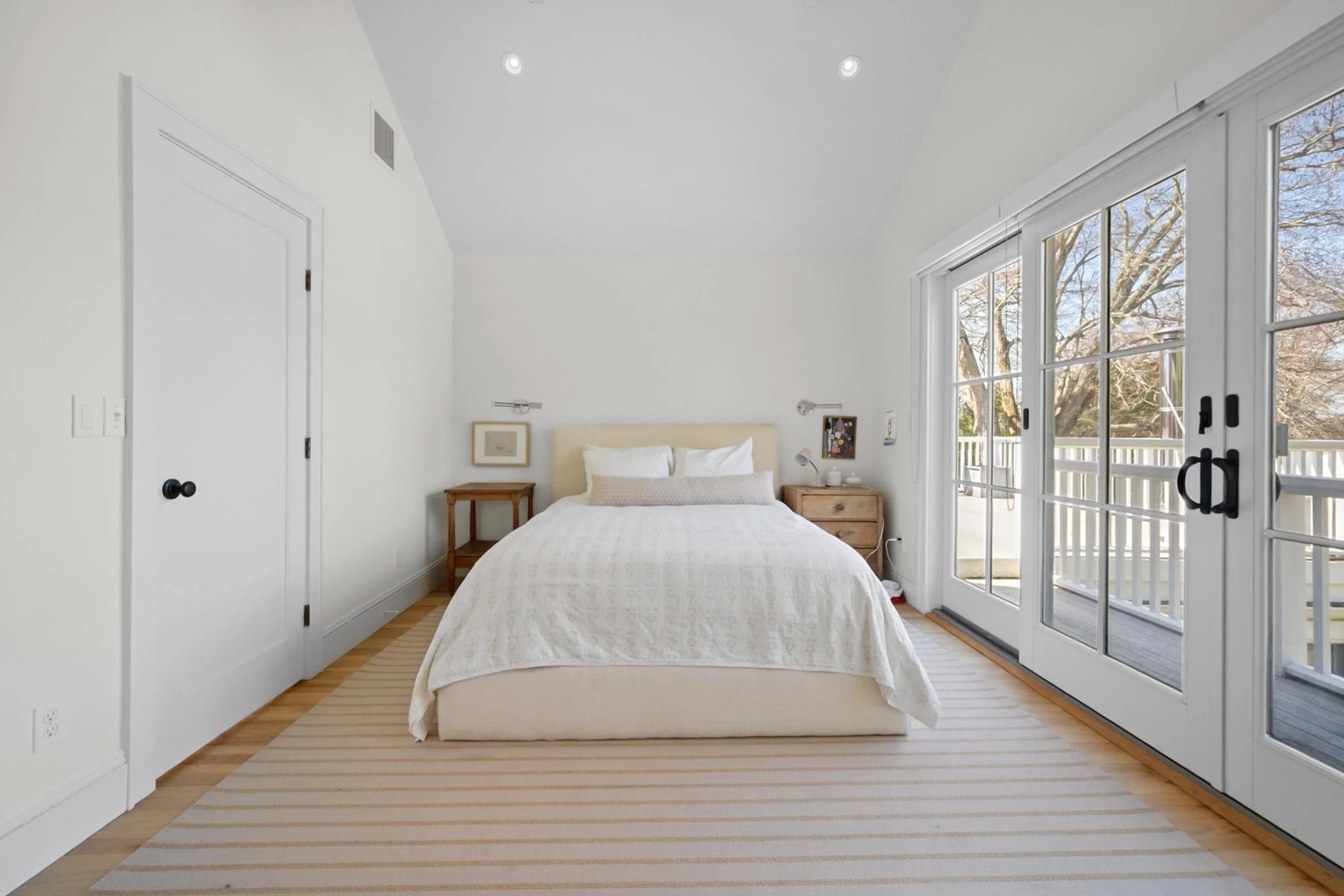 Bright, minimal bedroom with a white bed and linens, wood side tables, wall-mounted reading lights, framed art, and large glass sliding doors leading to a balcony with trees outside.