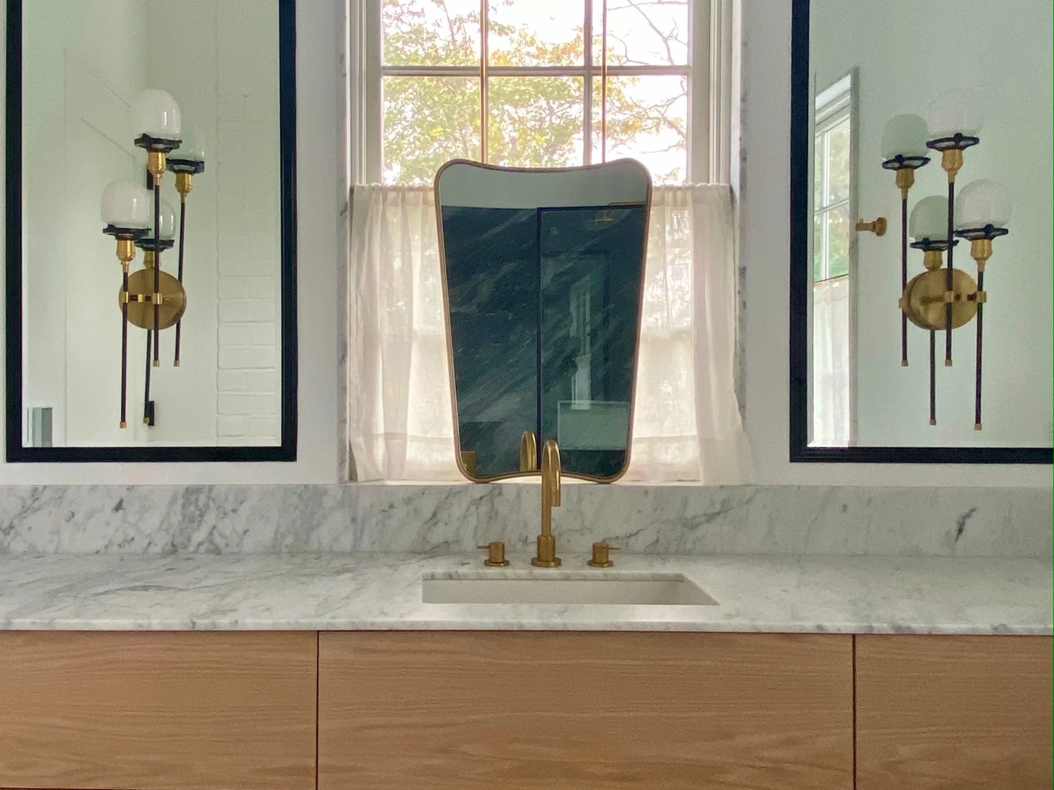 A bathroom vanity with a marble countertop, a gold faucet, and a mirror. On the left and right sides of the mirror are two black frames with artistic wall sconces that have two white glass bulbs each. Behind the mirror, a window allows natural light 