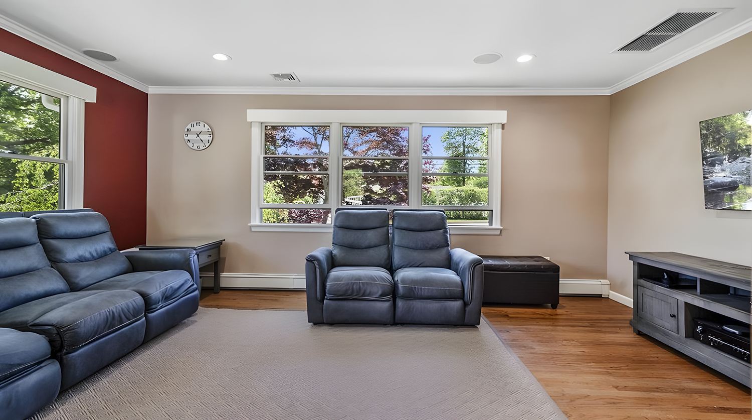 Living room with beige walls, red accent wall, hardwood flooring, large windows, black leather sofa, black leather armchair, gray TV stand, wall-mounted TV, wall clock, ceiling vents, and through the windows trees and greenery outside.