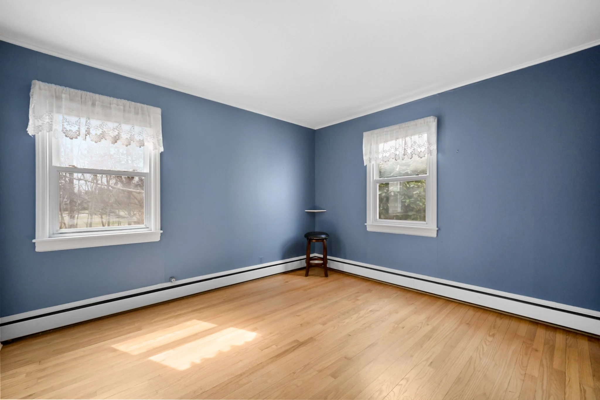 A room with blue walls, two windows with lace curtains, hardwood flooring, and a single chair.