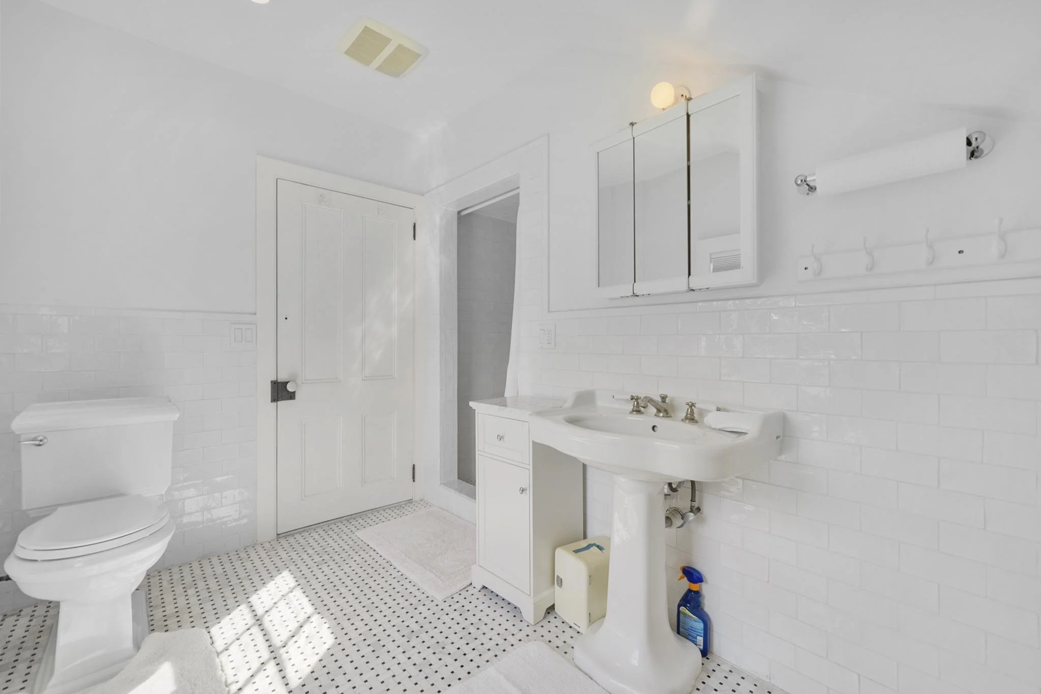 A white bathroom with a toilet, white vanity with sink, mirror cabinets, towel hooks, and a ceiling vent.