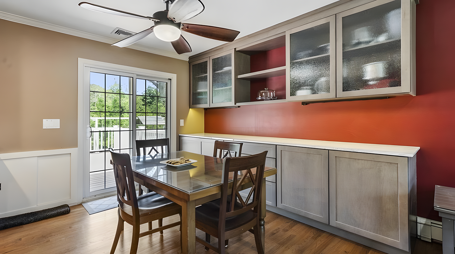 Dining room with wooden table and chairs, sliding glass door leading to a deck, kitchen cabinets with frosted glass doors, a ceiling fan, and an outside view of greenery.