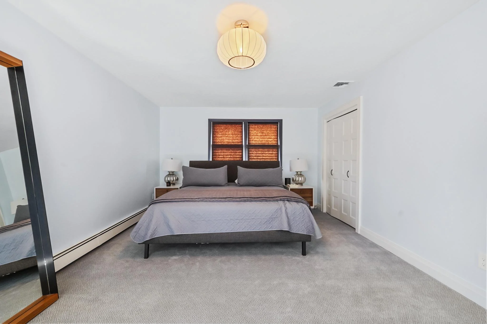 A bedroom with white walls, a bed with grey bedding and pillows, two matching nightstands with lamps, a large mirror, a window with blinds, a closet door, and a ceiling light fixture.