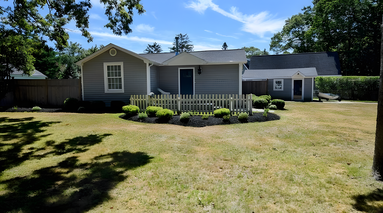 A backyard with a gray house, a small white picket fence, bushes, a boat, and a lawn with trees in the background under a blue sky.