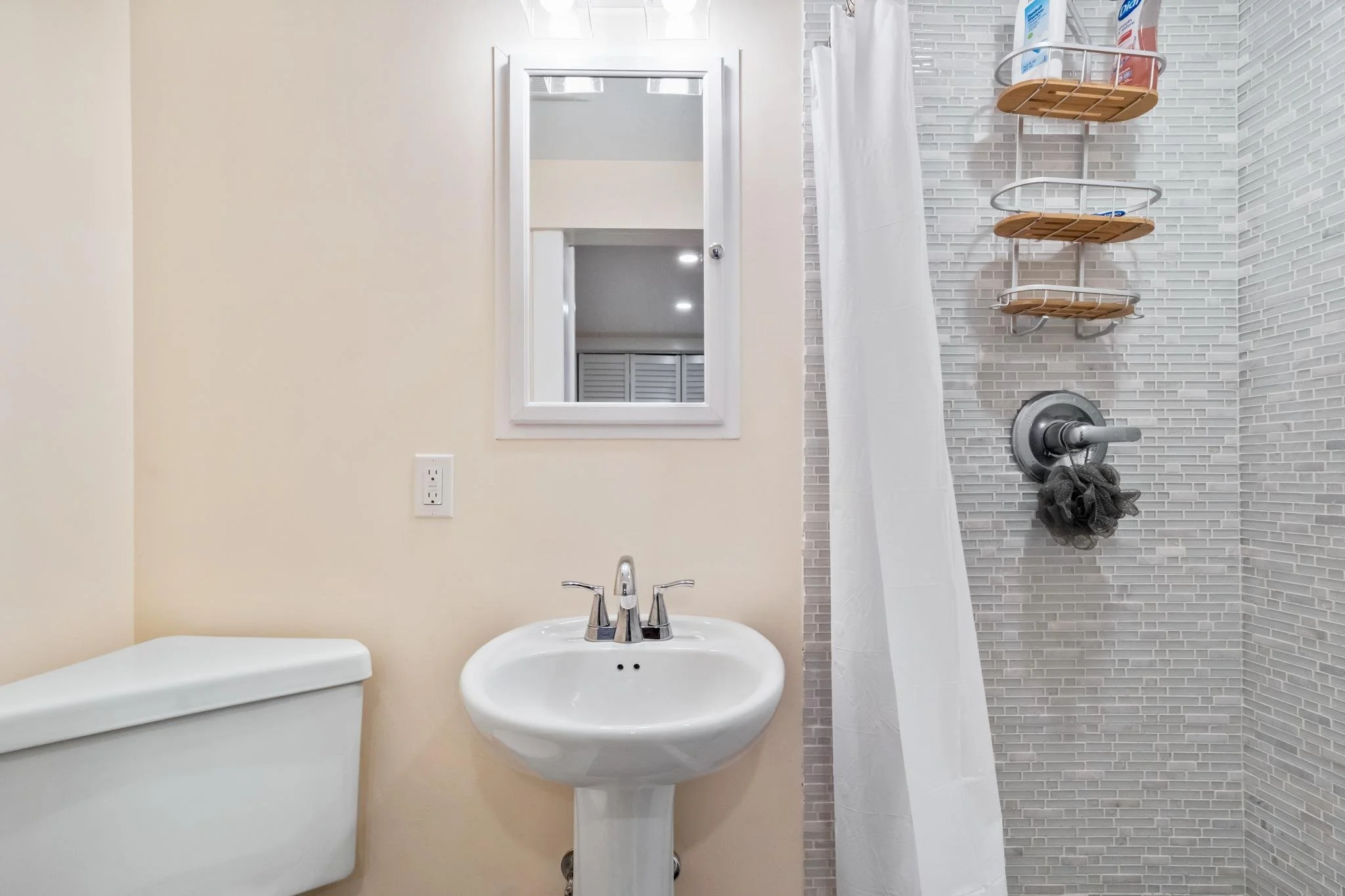 Bathroom with beige walls, a small mirror, a bathroom sink with a faucet, a white toilet, and a shower area with gray tile walls, a white shower curtain, a wooden and metal shower shelf with toiletries, and a gray bath pouf hanging from the shower ha