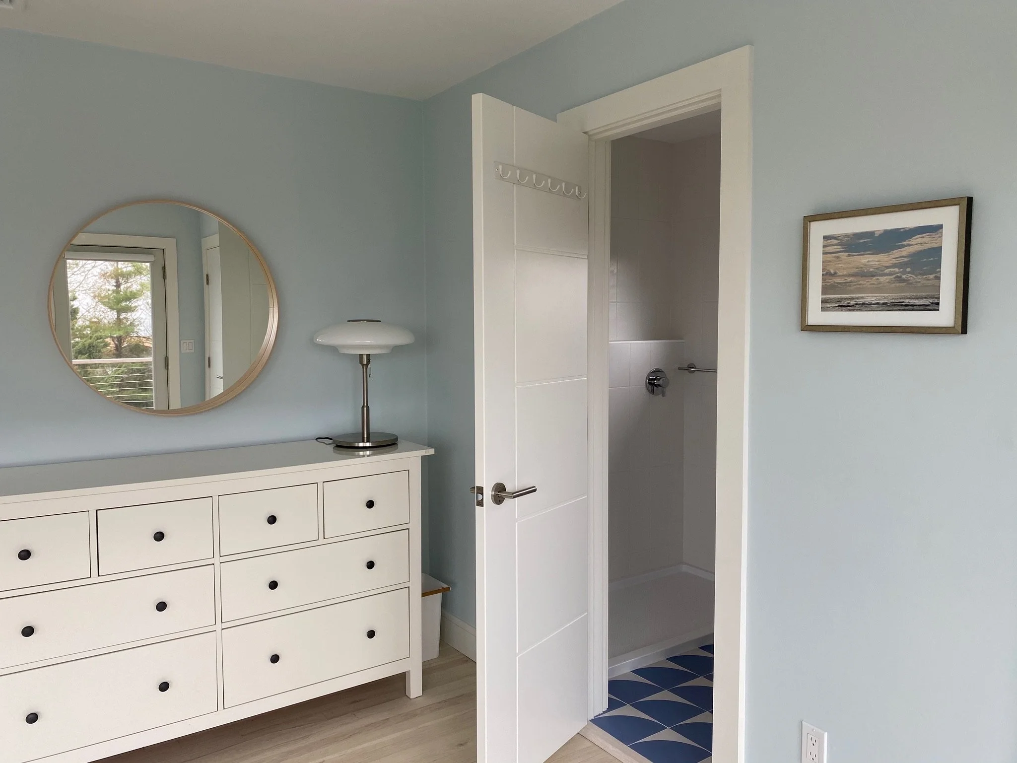 A bathroom with a white dresser with black knobs, a circular mirror, a window showing trees outside, a standing lamp, and a bathroom seen through an open door with towel hooks and tiled walls. There is also a framed picture of a landscape on the ligh