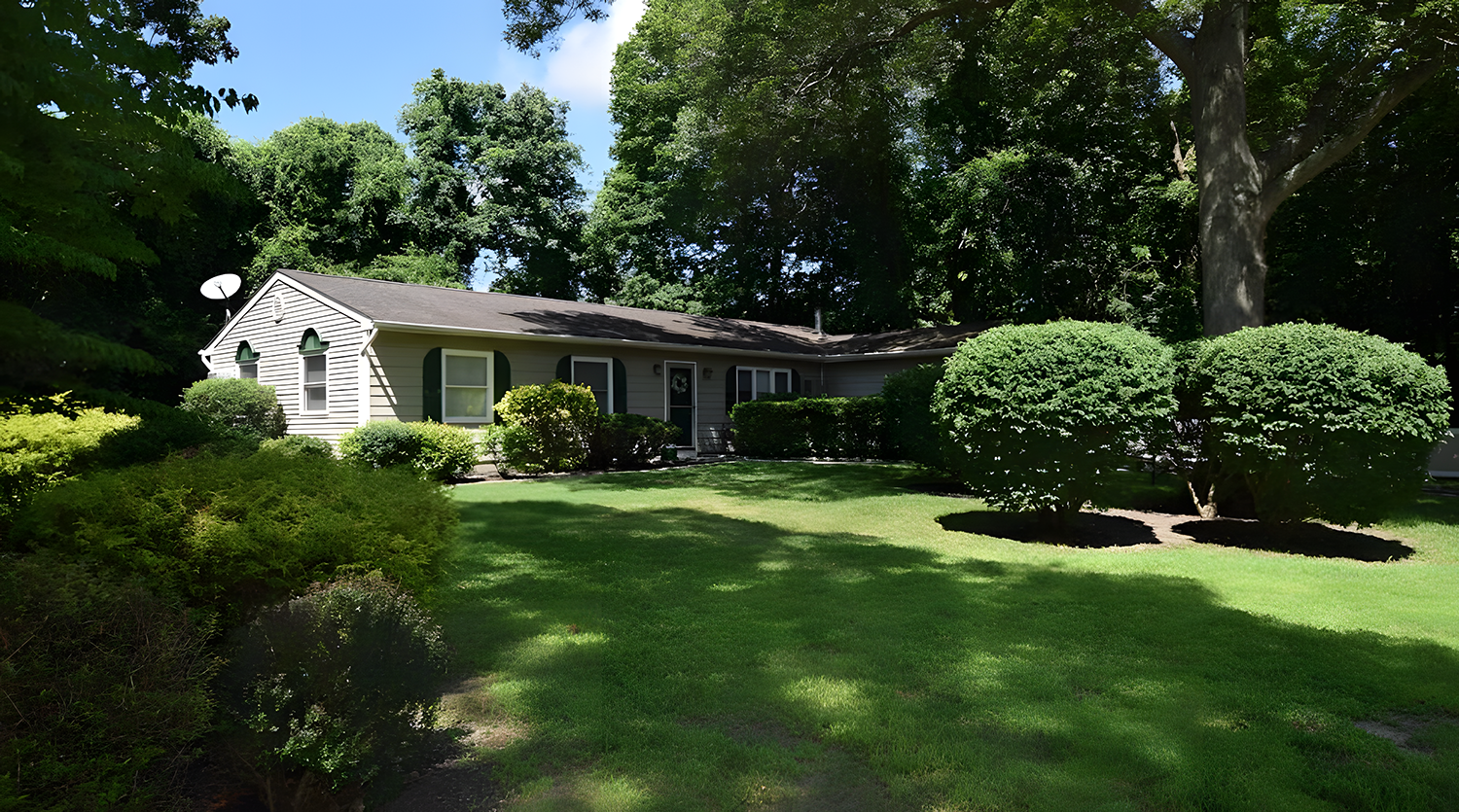 Single-story house with grey siding, black shutters, and an attached small porch, surrounded by lush green bushes and trees in a well-maintained yard, with sunlight and blue sky in the background.