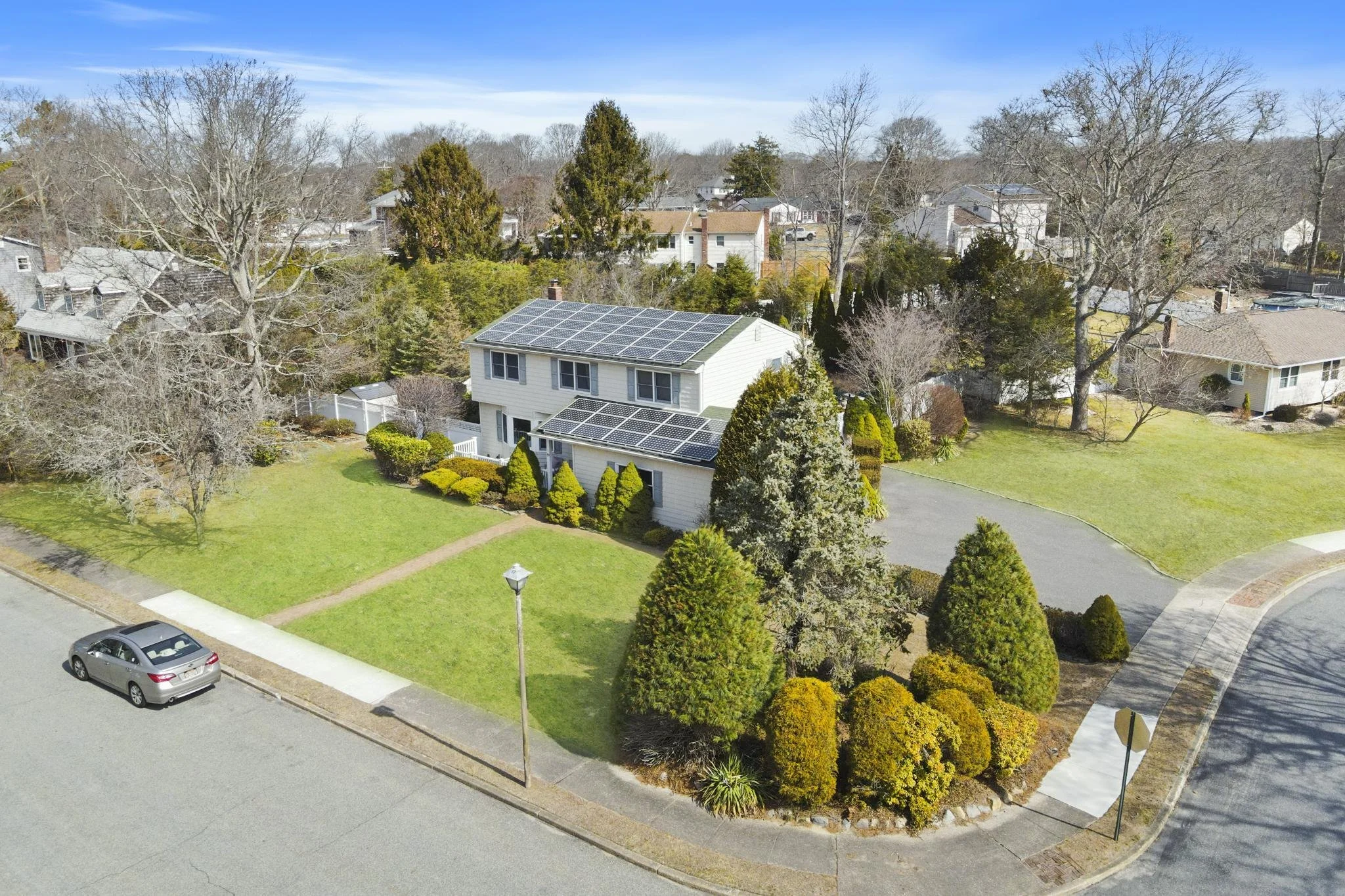 Aerial view of a suburban neighborhood with houses, trees, a well-maintained lawn, a car parked on the street, and solar panels on the roof of a white house.