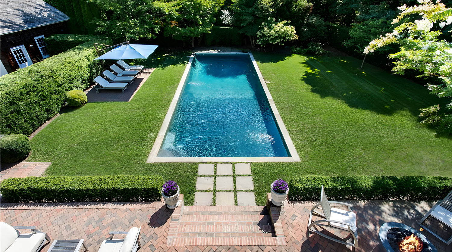 A backyard with a rectangular swimming pool surrounded by a grassy lawn, garden hedges, and trees. There are lounge chairs with a large umbrella on a patio area, and a brick pathway leading to the pool. Two flower pots with purple flowers are on eith