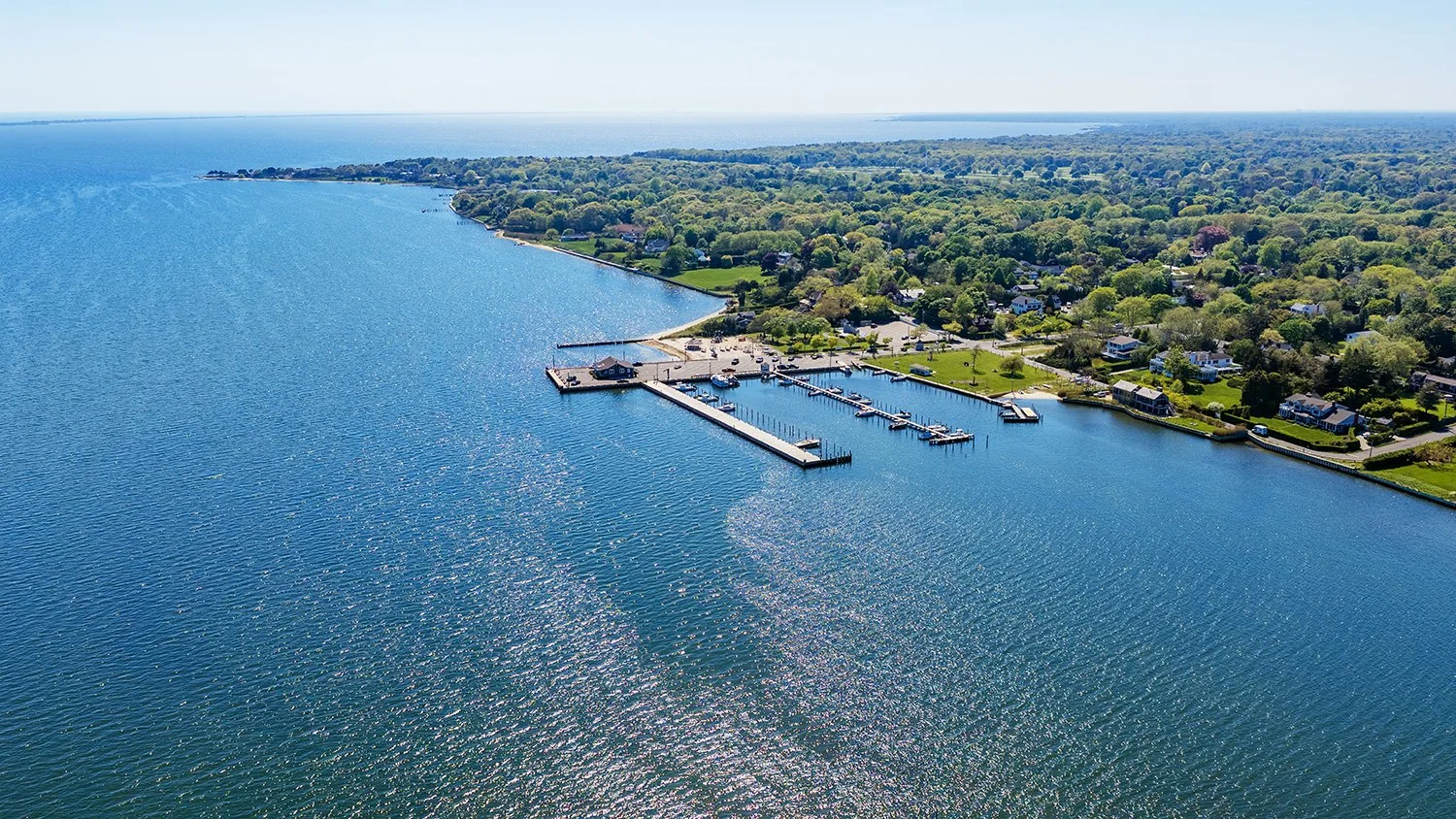 Aerial view of a marina with boats docked along piers, adjacent to a residential neighborhood with trees and houses, next to a large body of water under a clear sky.
