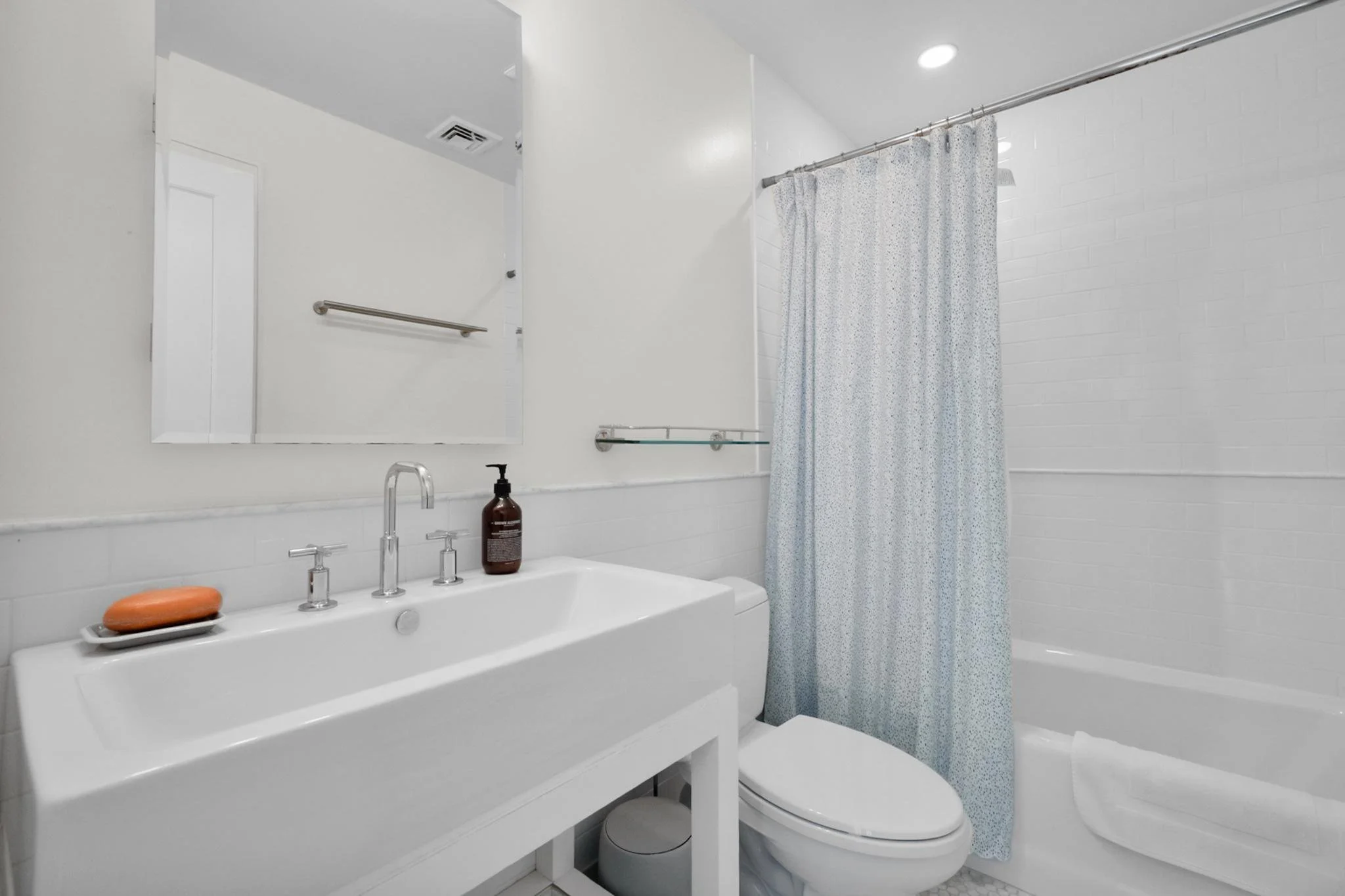 A modern, minimalistic bathroom with a white sink, a mirror, a soap dispenser, a shower with a patterned curtain, and a toilet, all with a white and light grey color scheme.