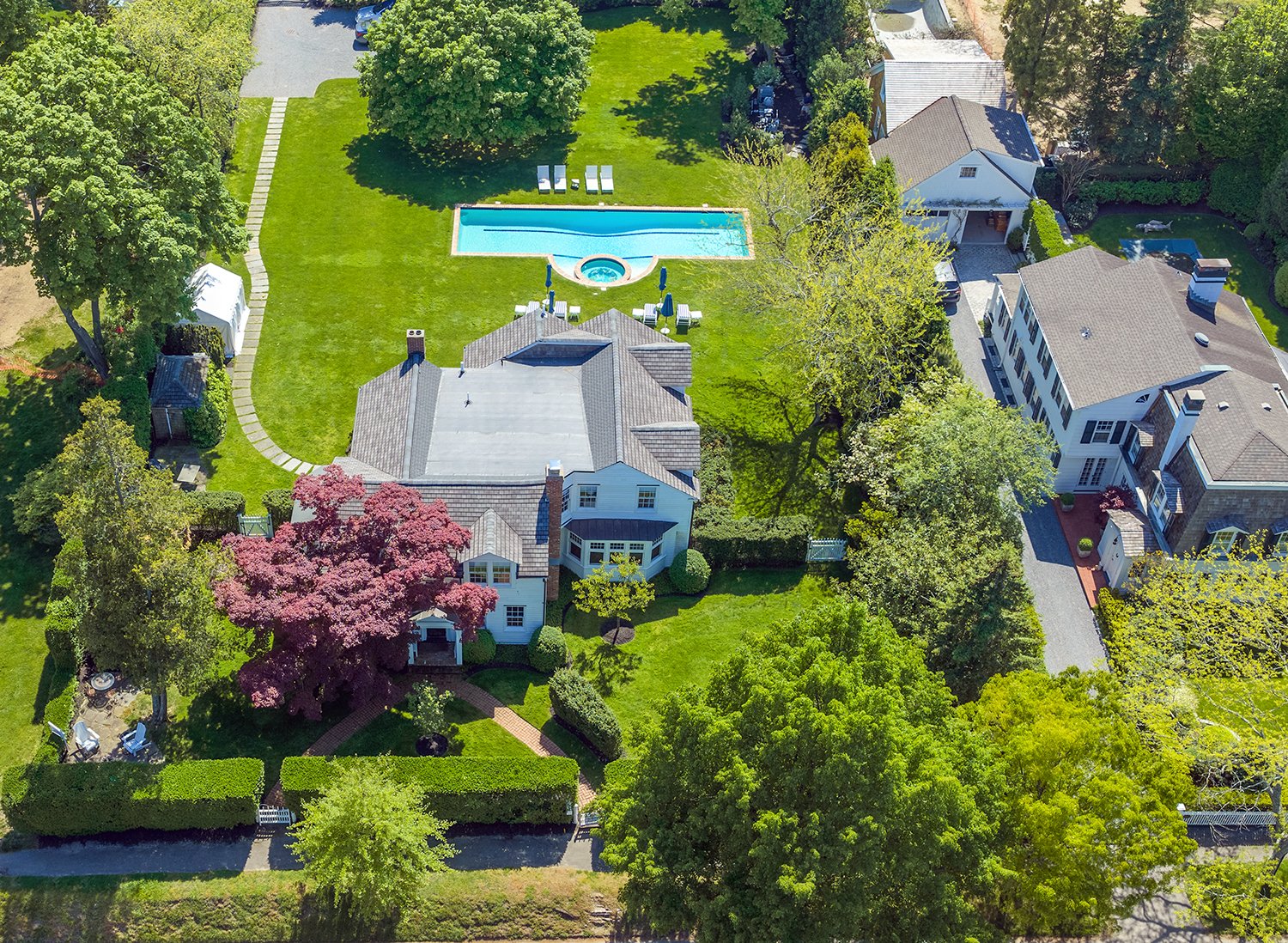 Aerial view of a backyard with a swimming pool, surrounded by green trees, a manicured lawn, and two houses with outdoor sitting areas and pathways.