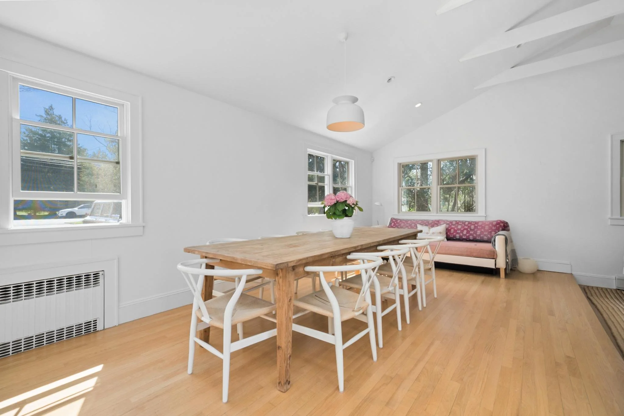 Bright dining room with a wooden table surrounded by white chairs, a pink floral-patterned bench, a vase with pink flowers, and multiple windows letting in sunlight.