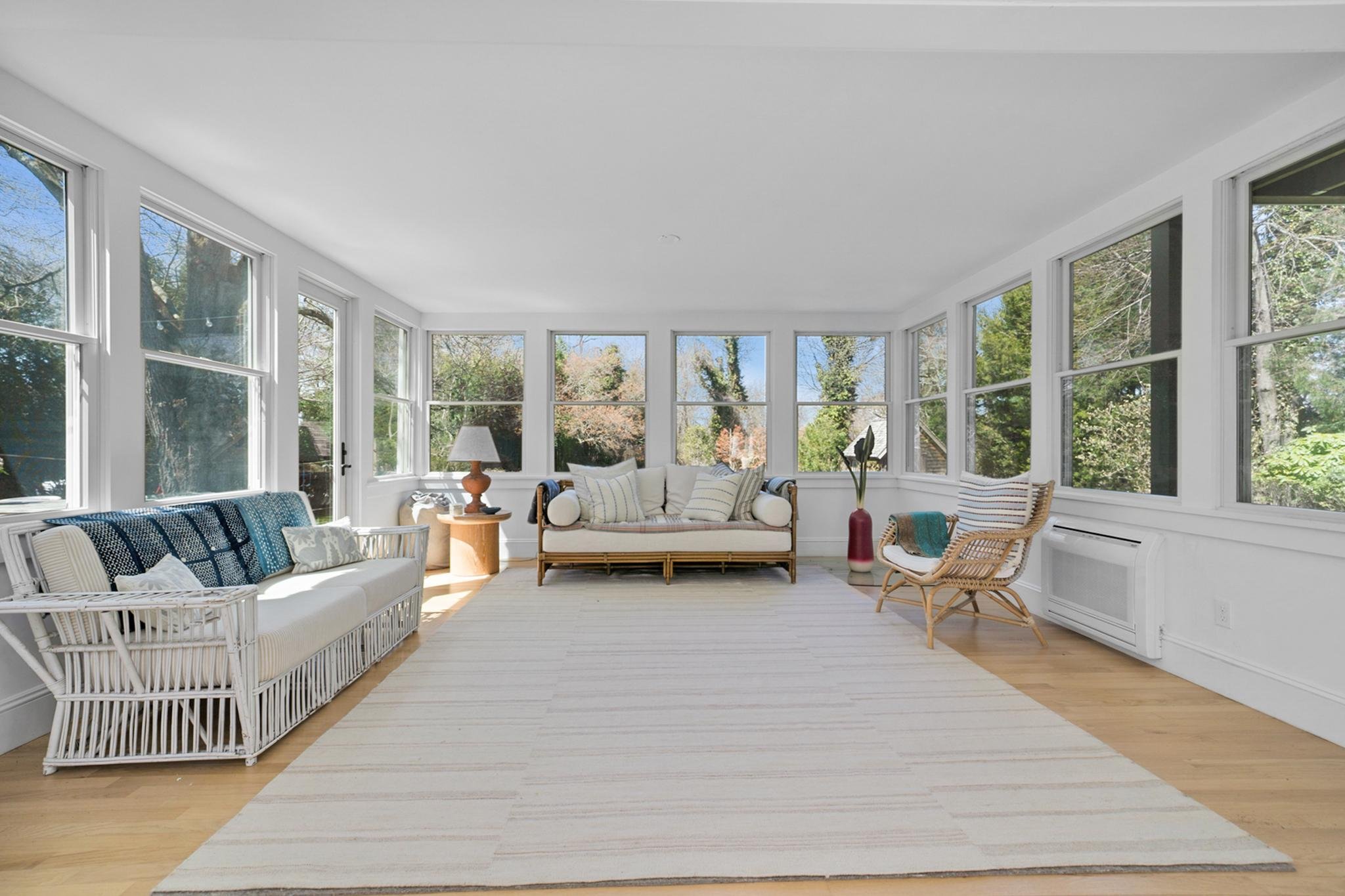 A bright sunlit living room with large windows showing trees outside, featuring white walls, a white rug, wicker and upholstered furniture, a wooden side table with a lamp, and various pillows and decorations.