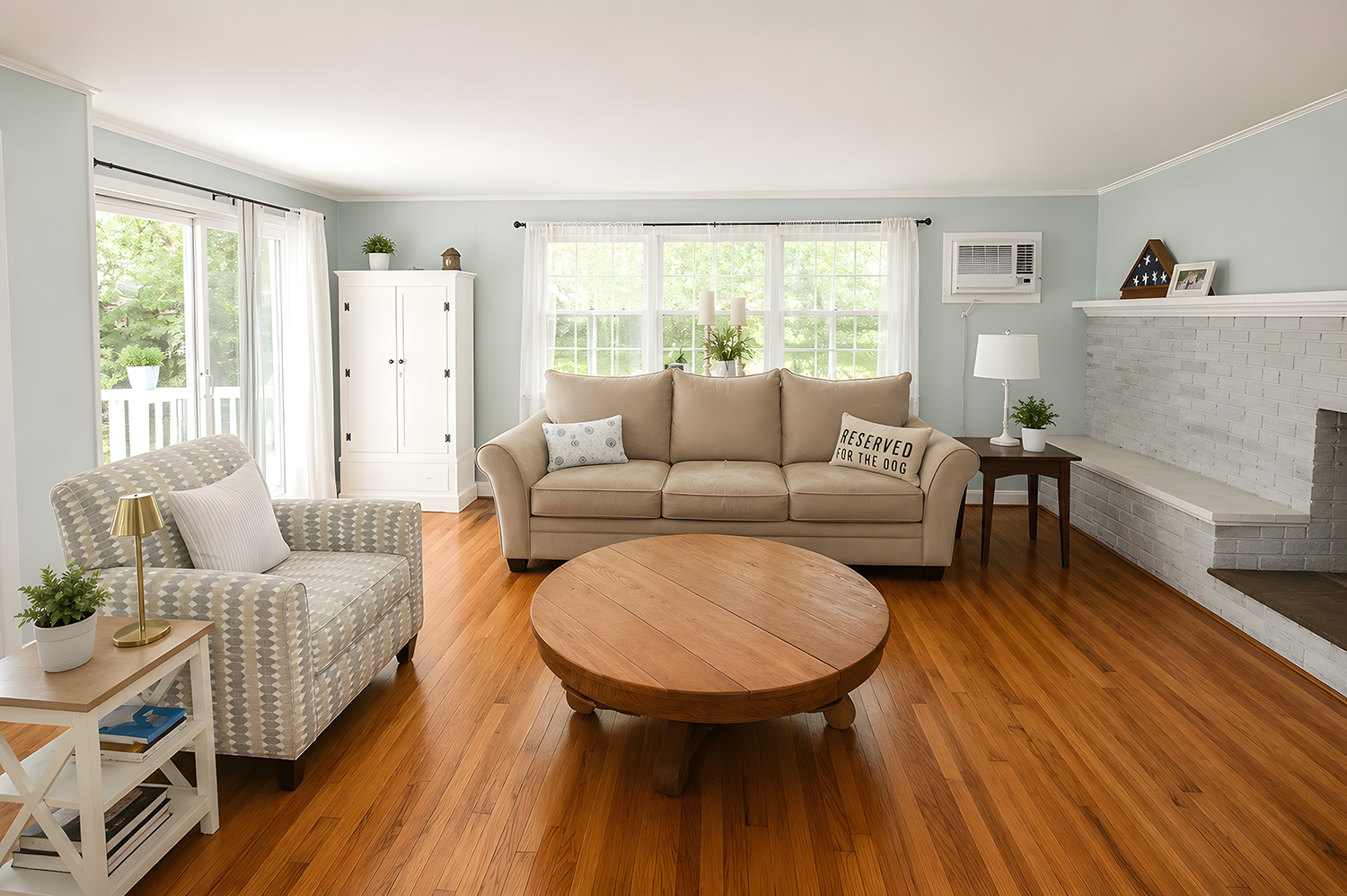 Bright living room with beige sofa, armchair, wooden coffee table, white fireplace, and large windows with white curtains, decorated with plants and picture frames.