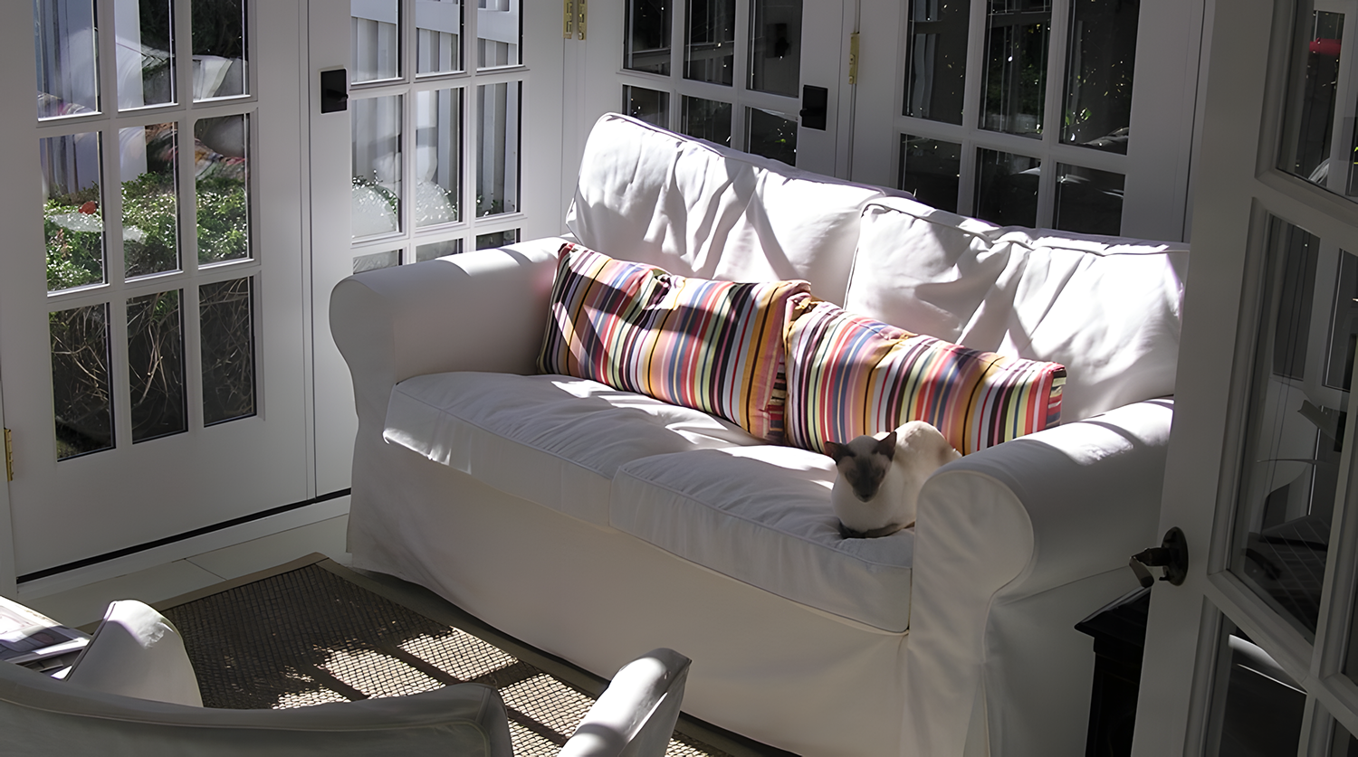 Sunlit white sofa with a colorful striped pillow and a Siamese cat resting on it in a sunroom with large glass windows.