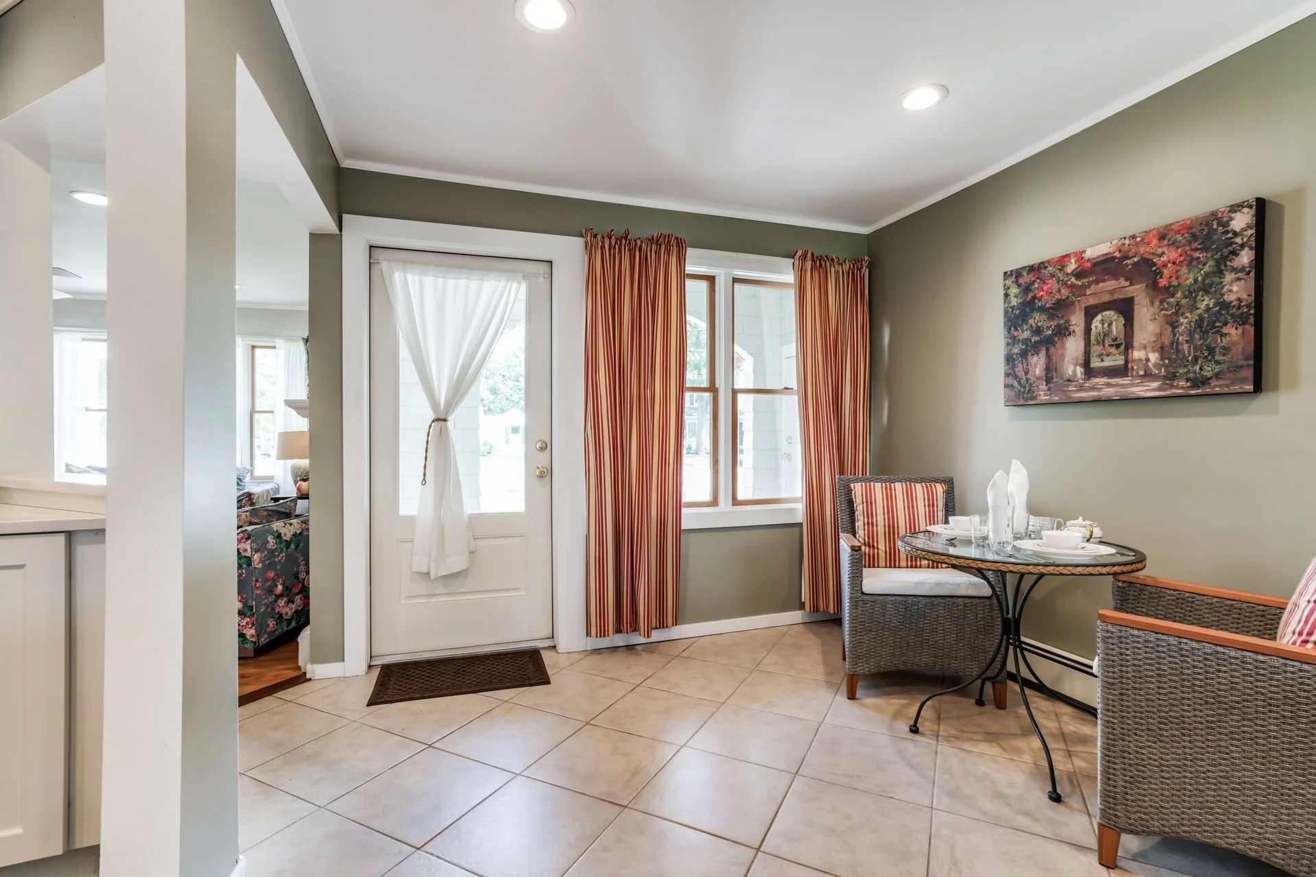 A cozy dining area with a round table set with white tableware, surrounded by wicker chairs with striped cushions. The light green walls feature a large landscape painting, and there are windows with red and white striped curtains. A glass door with 