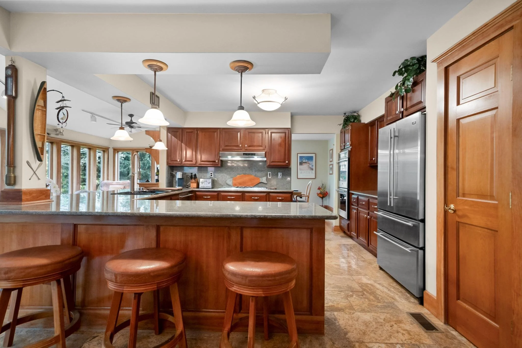 Kitchen with wooden cabinets, stainless steel appliances, granite island, three bar stools, and pendant lighting.