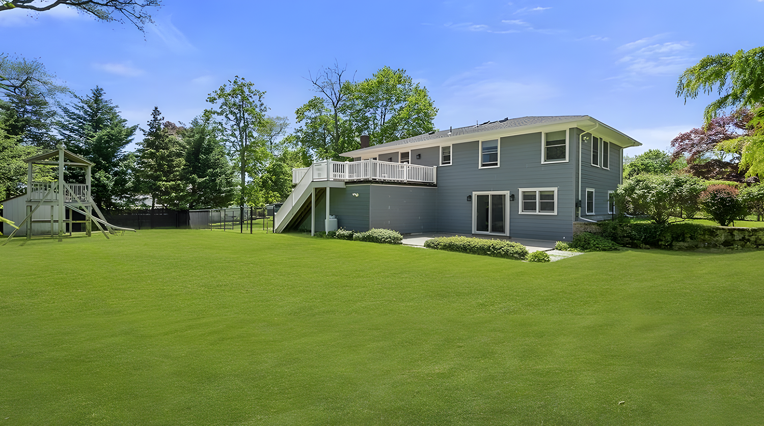 A blue two-story house with a large backyard, green grass, trees, and a wooden children’s play structure with a slide.
