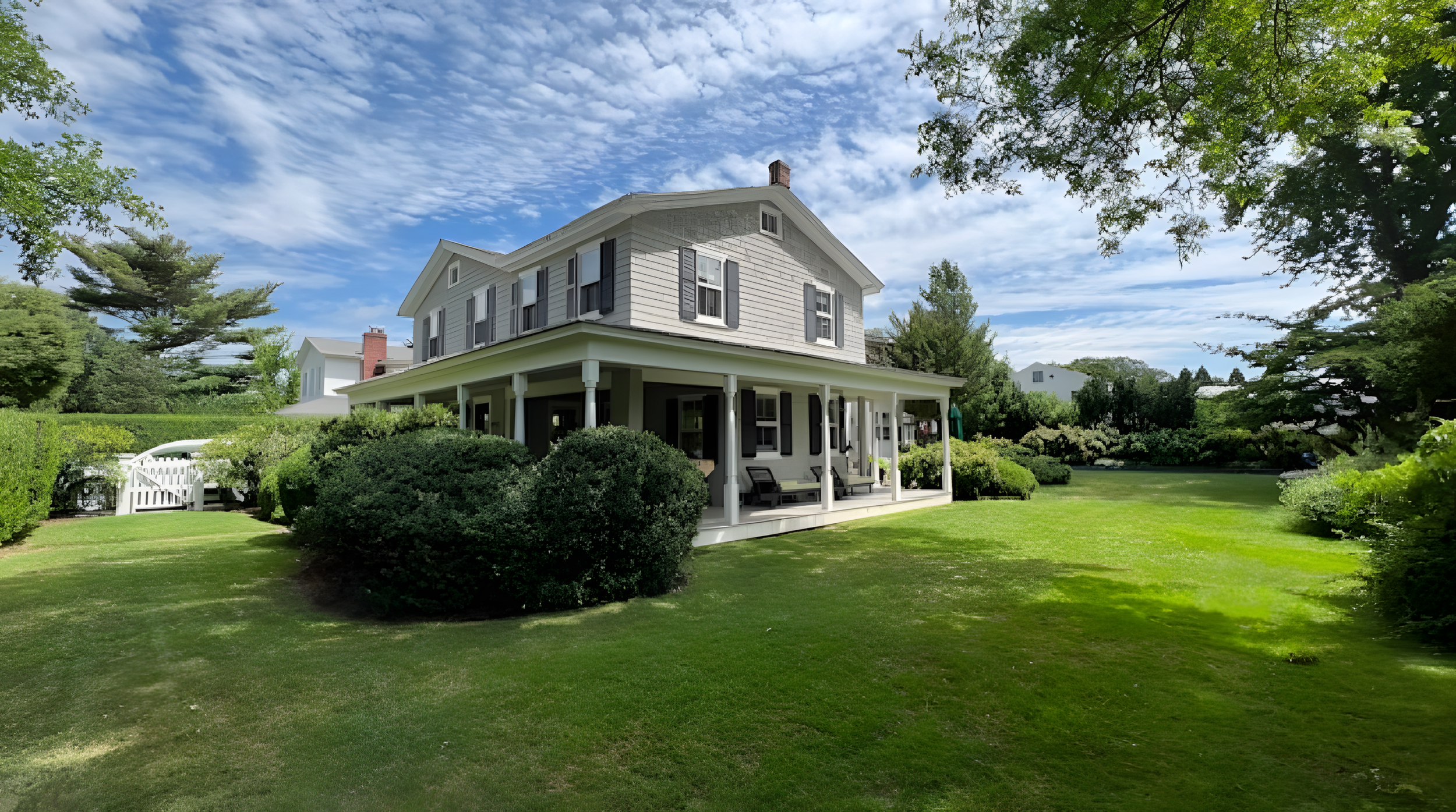 White two-story house with a large front porch, surrounded by well-maintained green lawn and trees on a sunny day with a partly cloudy sky.