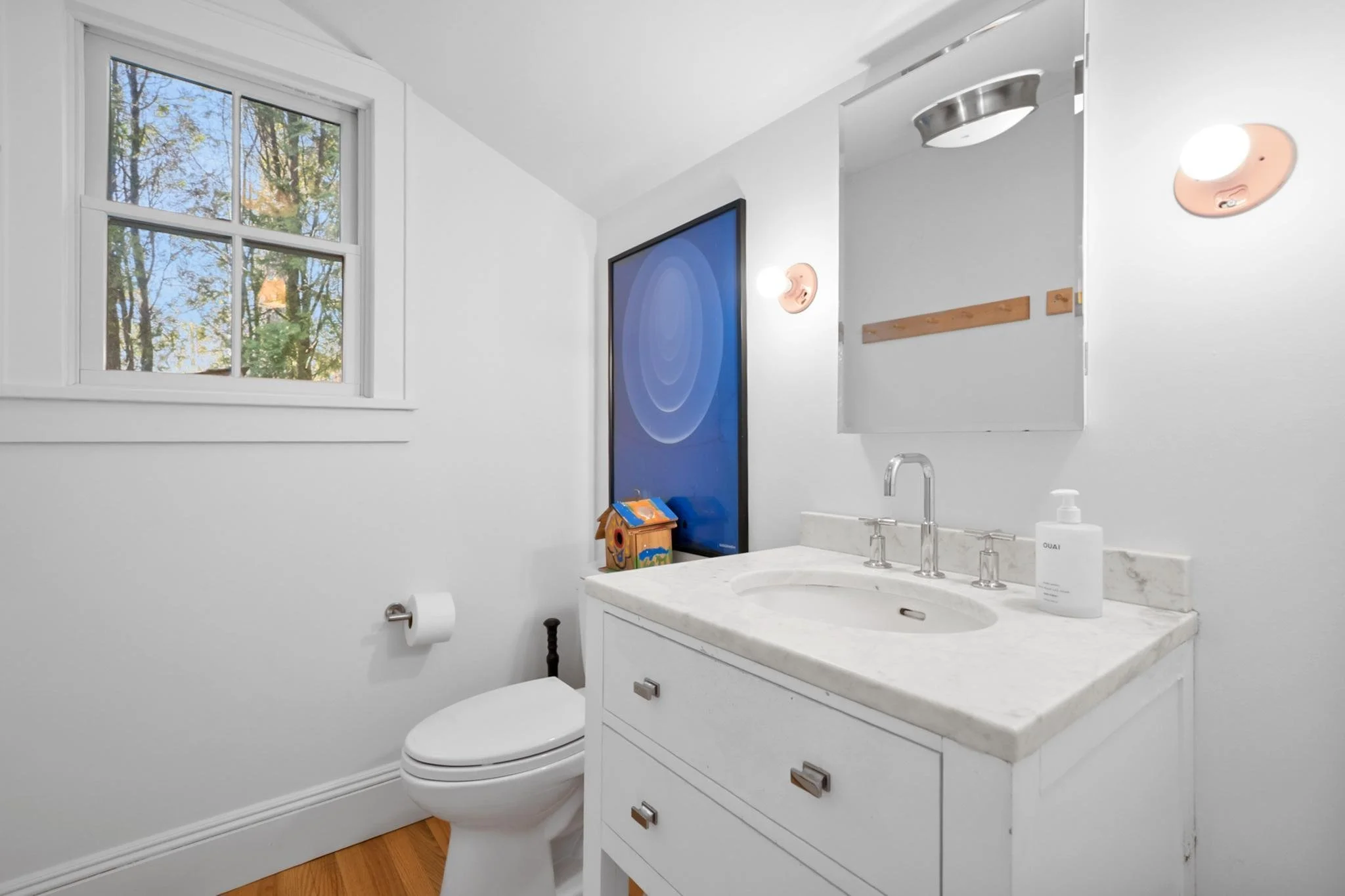 A small bathroom with a white vanity, marble countertop, and a sink with a chrome faucet. There is a mirror on the wall with two small light fixtures on either side. A window shows trees outside, allowing natural light into the room. The toilet paper