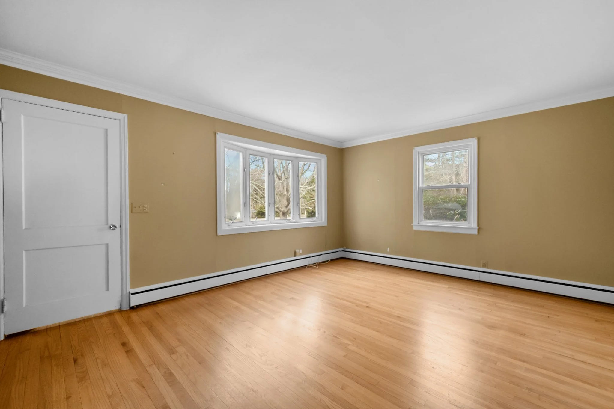 Empty room with beige walls, white trim, hardwood floors, and two large windows letting in natural light.