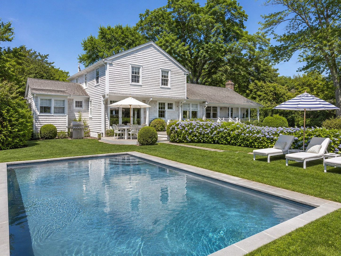 A backyard with a swimming pool, green lawn, white house with a patio, umbrellas, lounge chairs, and blooming flowers amid trees on a sunny day.