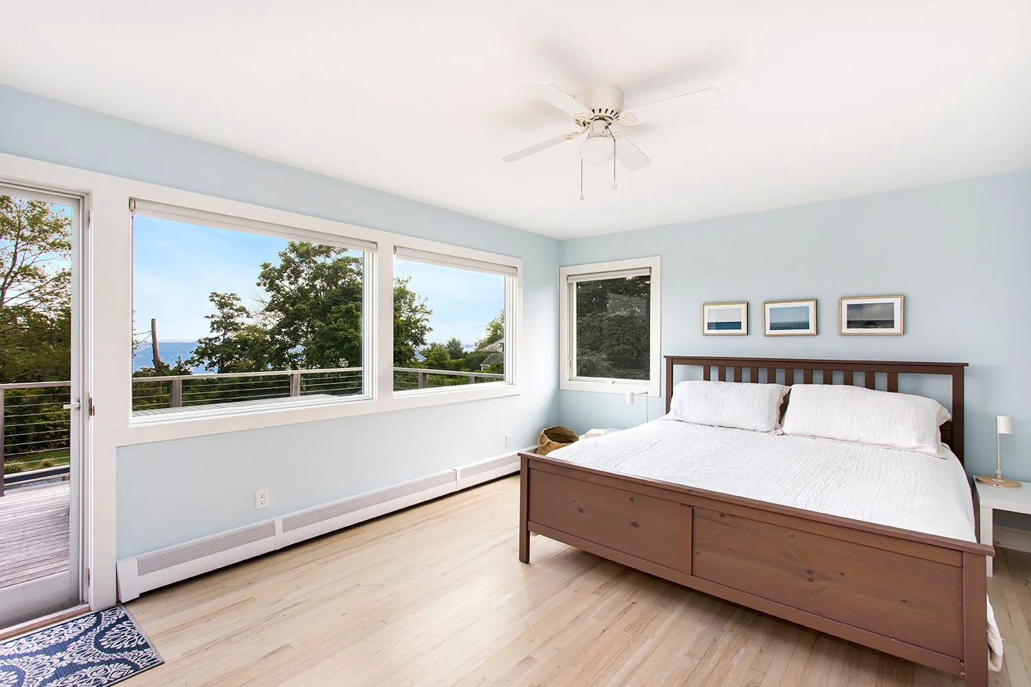 A bright bedroom with light blue walls, large windows showing trees and an outdoor deck, a wooden bed frame with white bedding, three framed pictures on the wall above the bed, and a ceiling fan.