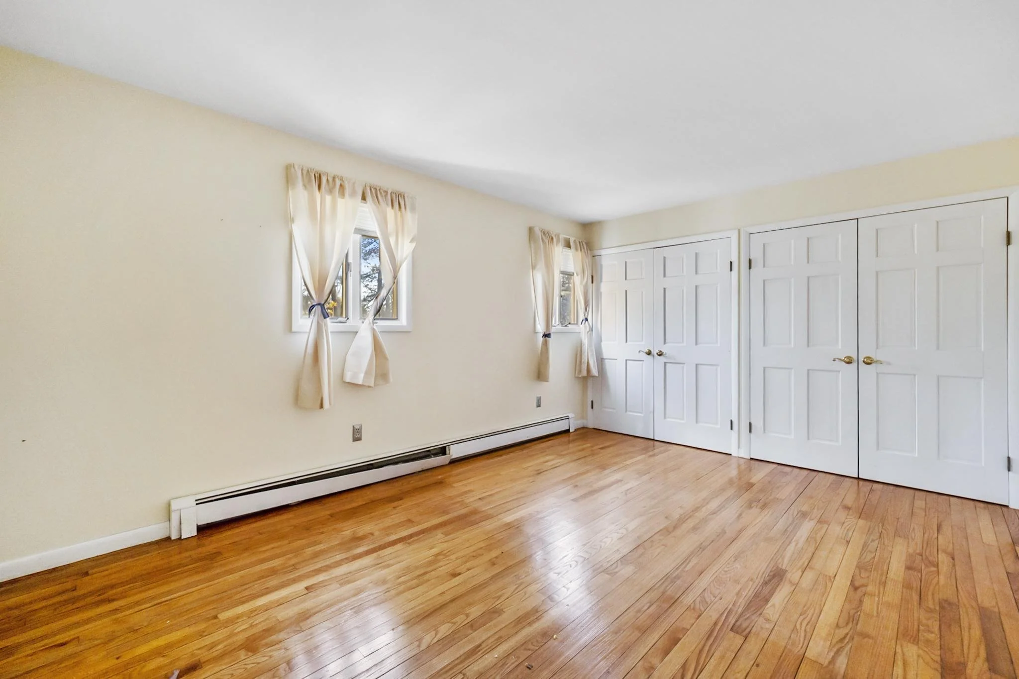 Empty room with hardwood floors, cream-colored walls, two windows with light curtains, a closet with double white doors, baseboard heater, and no furniture.
