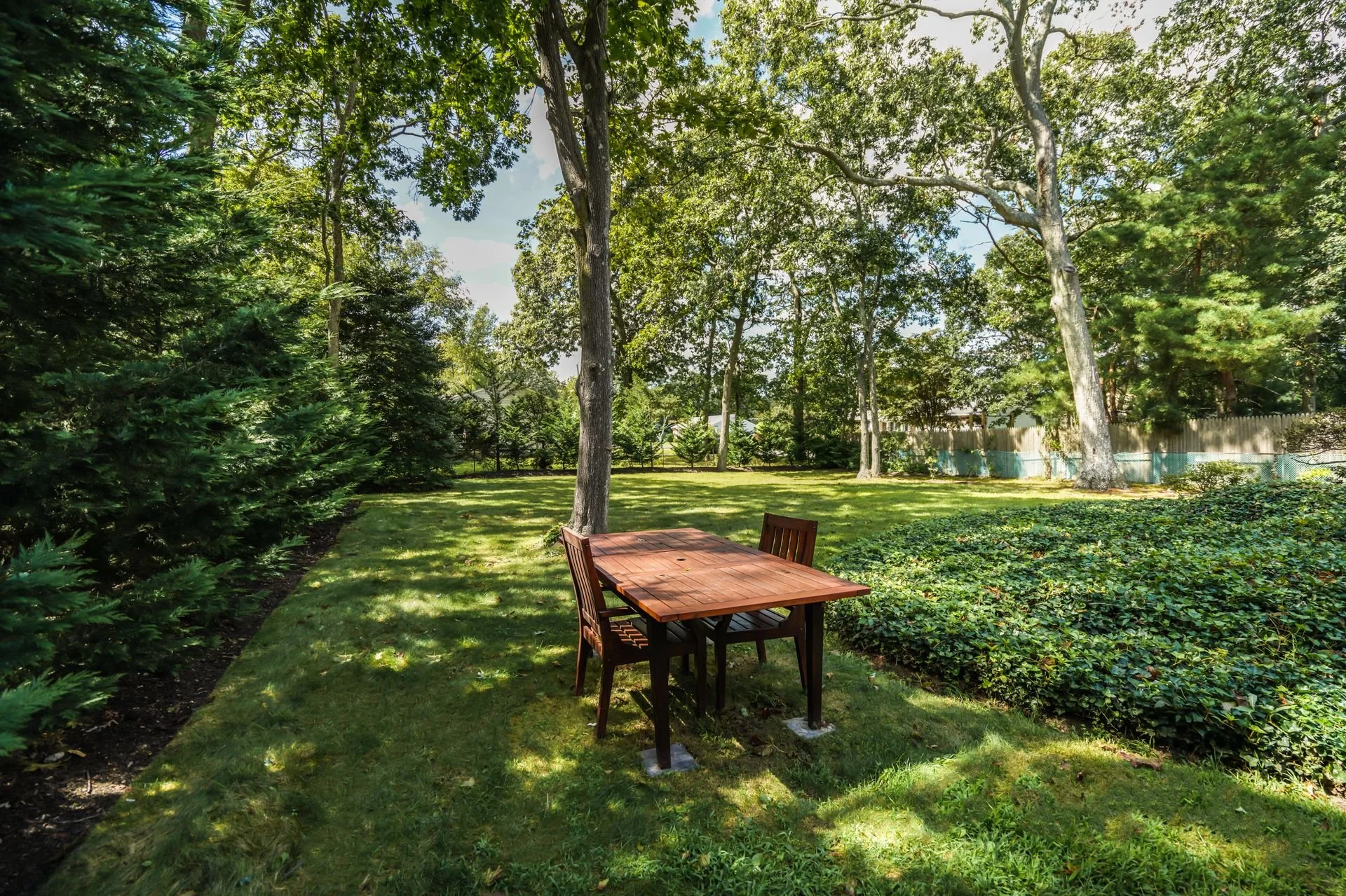 A backyard garden with a wooden table and chairs, surrounded by trees and lush greenery, on a sunny day.